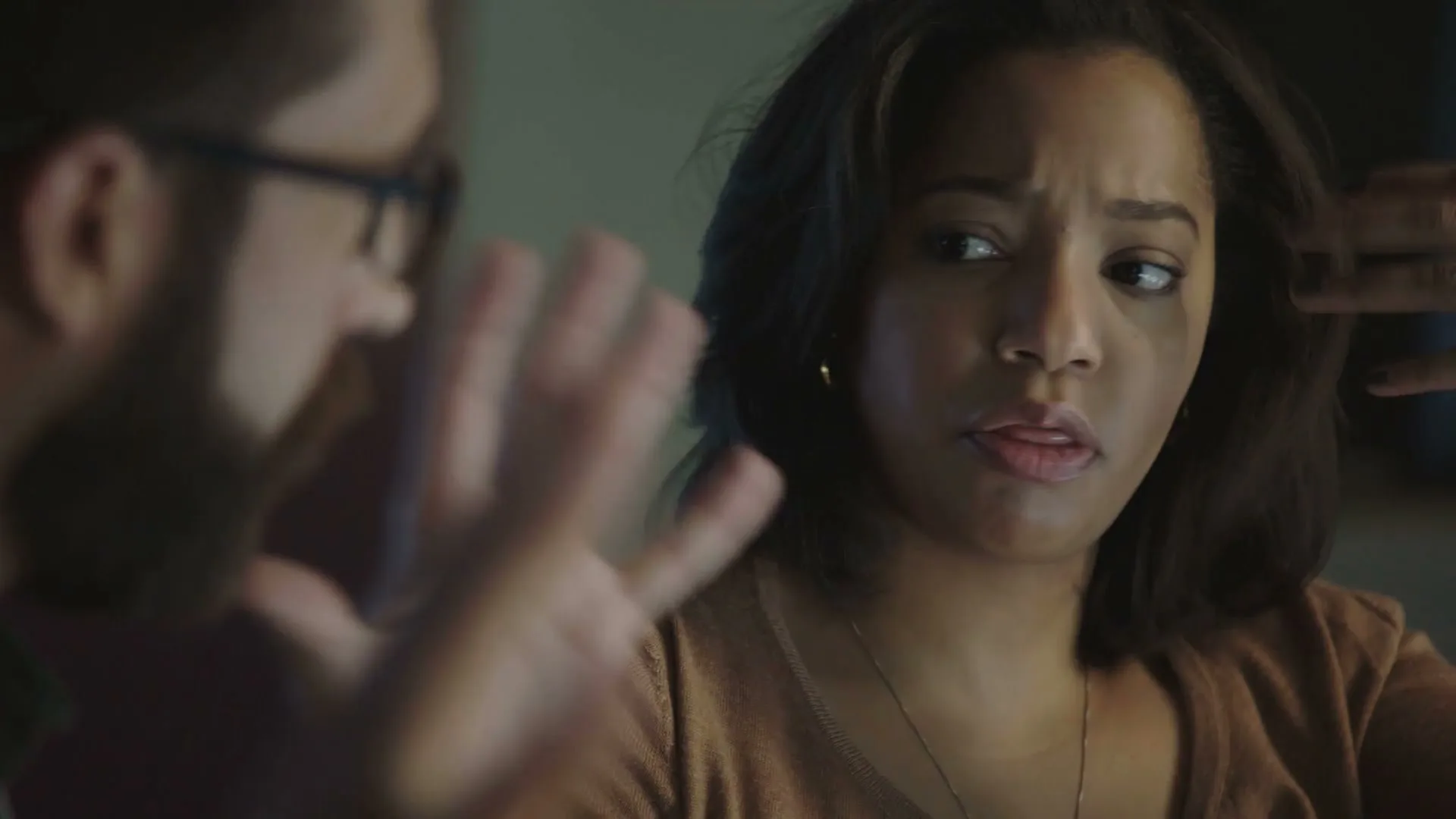 A young woman with dark hair and a brown top looks concerned while a bearded man in glasses gestures beside her, in an interior setting.