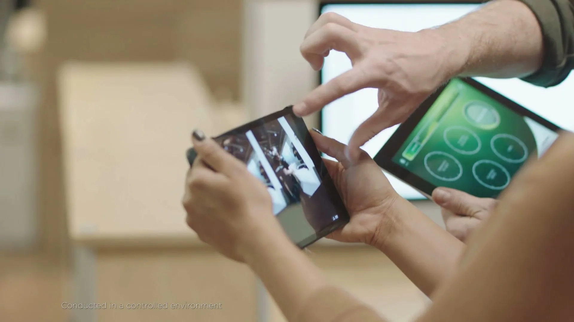 Two people's hands hold up two tablets, one showing a dark room with bright lights, the other a green interface, in an interior setting.