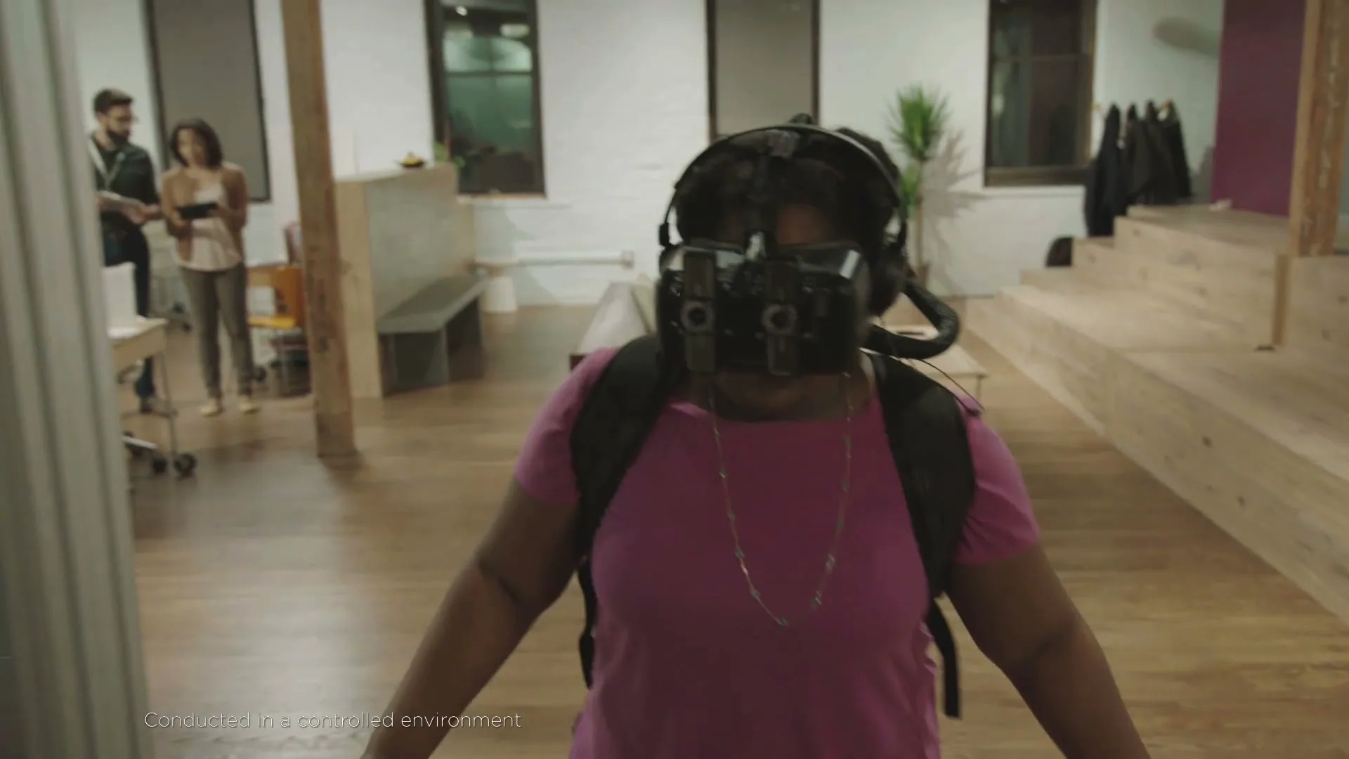 A woman wearing a VR headset and backpack walks forward in an open-plan office, while two people observe her in the background.