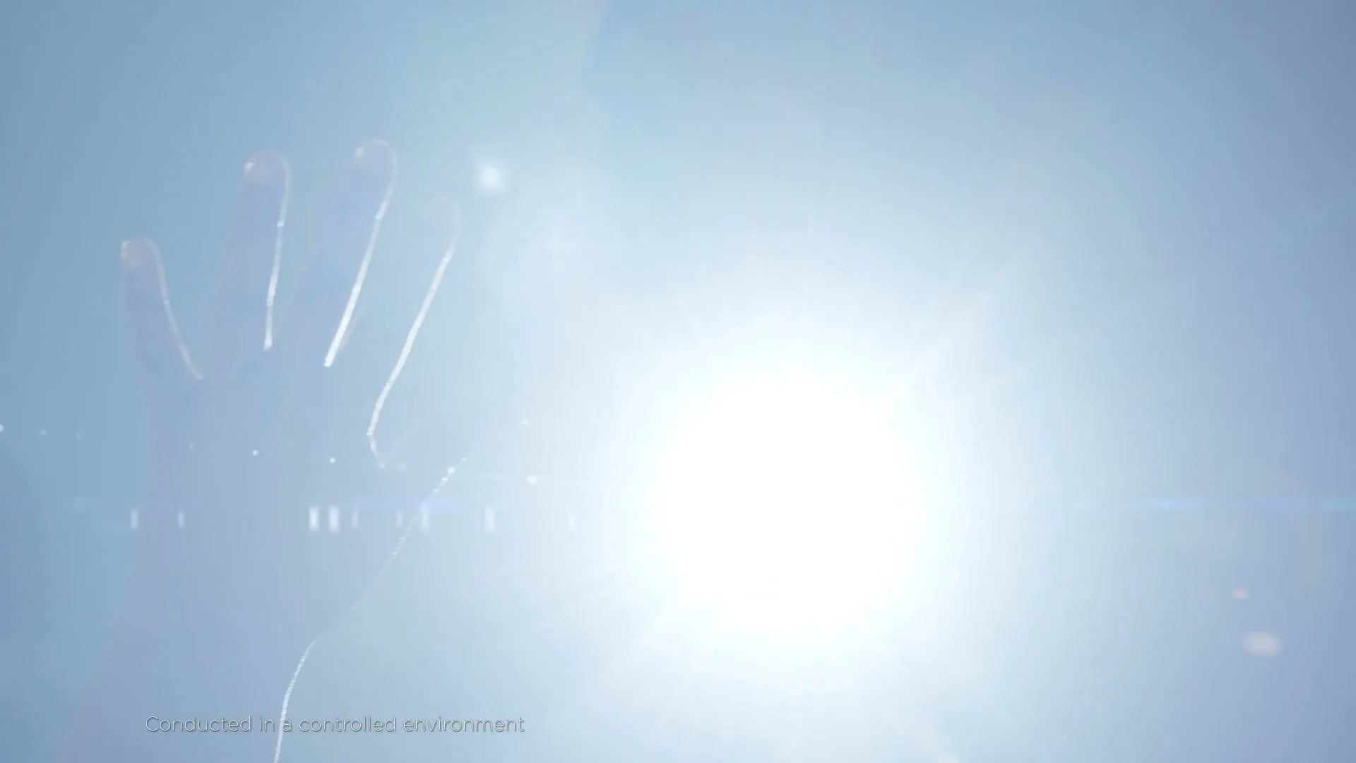 A person's hand is silhouetted against a bright, glaring light source, creating a lens flare effect in a controlled environment.