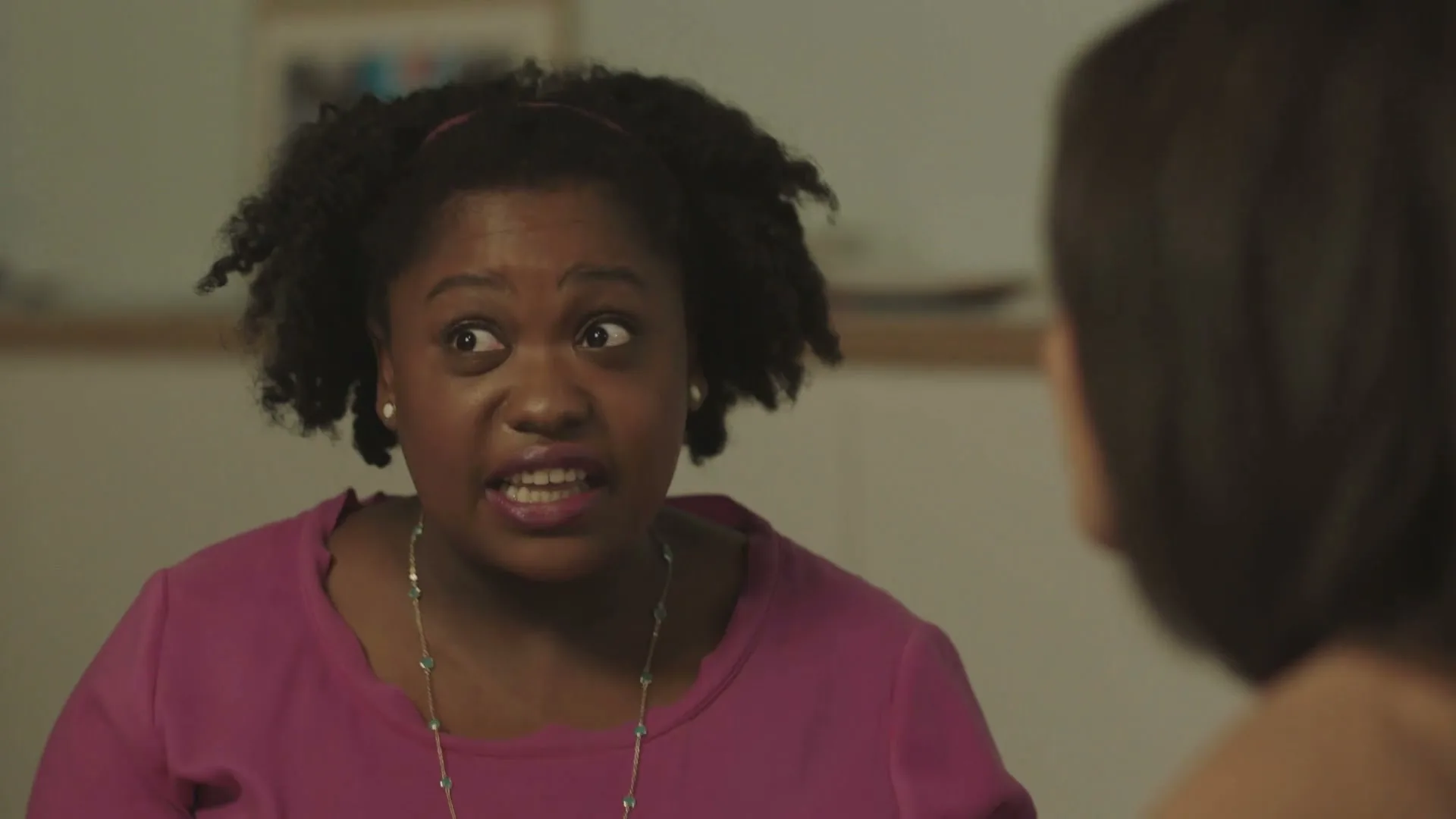 A young Black woman with curly hair and a pink top talks to someone off-screen, her expression shifting from calm to surprised and intense.