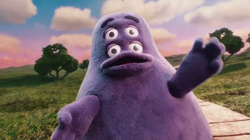 A purple, four-eyed, blob-like character with a surprised expression waves its hand in a whimsical, grassy landscape under a cloudy sky.