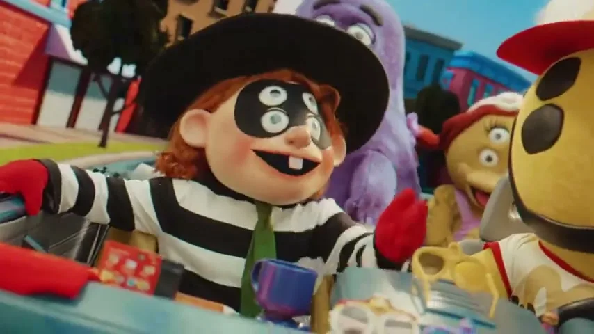 A puppet of the Hamburglar, with four eyes, sits in a car with other McDonald's characters, smiling and looking around.