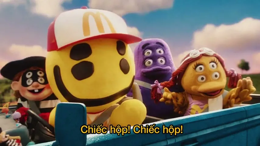 Four stylized, multi-eyed characters, including a large yellow smiley face in a McDonald's hat, ride in a blue vehicle on a sunny day.