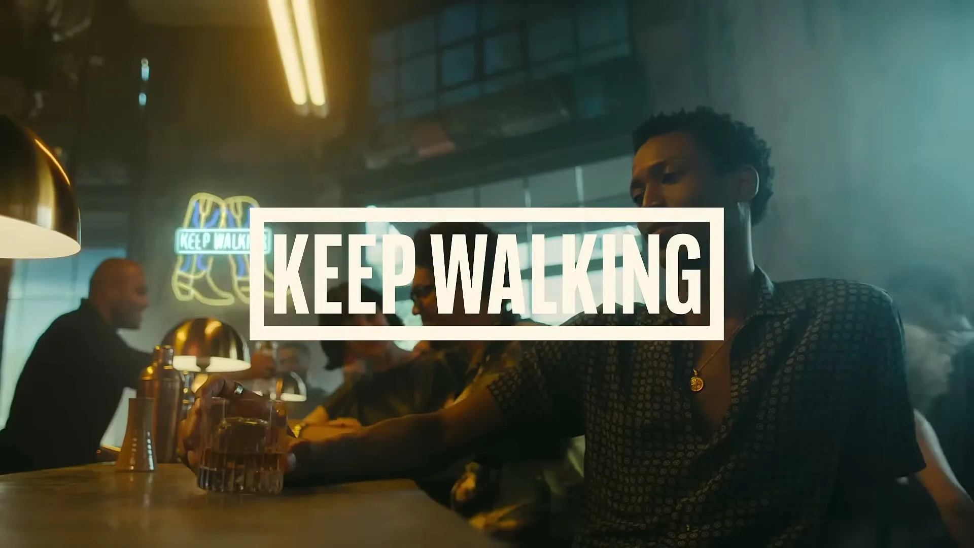 A man with dark curly hair sits at a bar with a drink, looking down, with "KEEP WALKING" text overlaid.