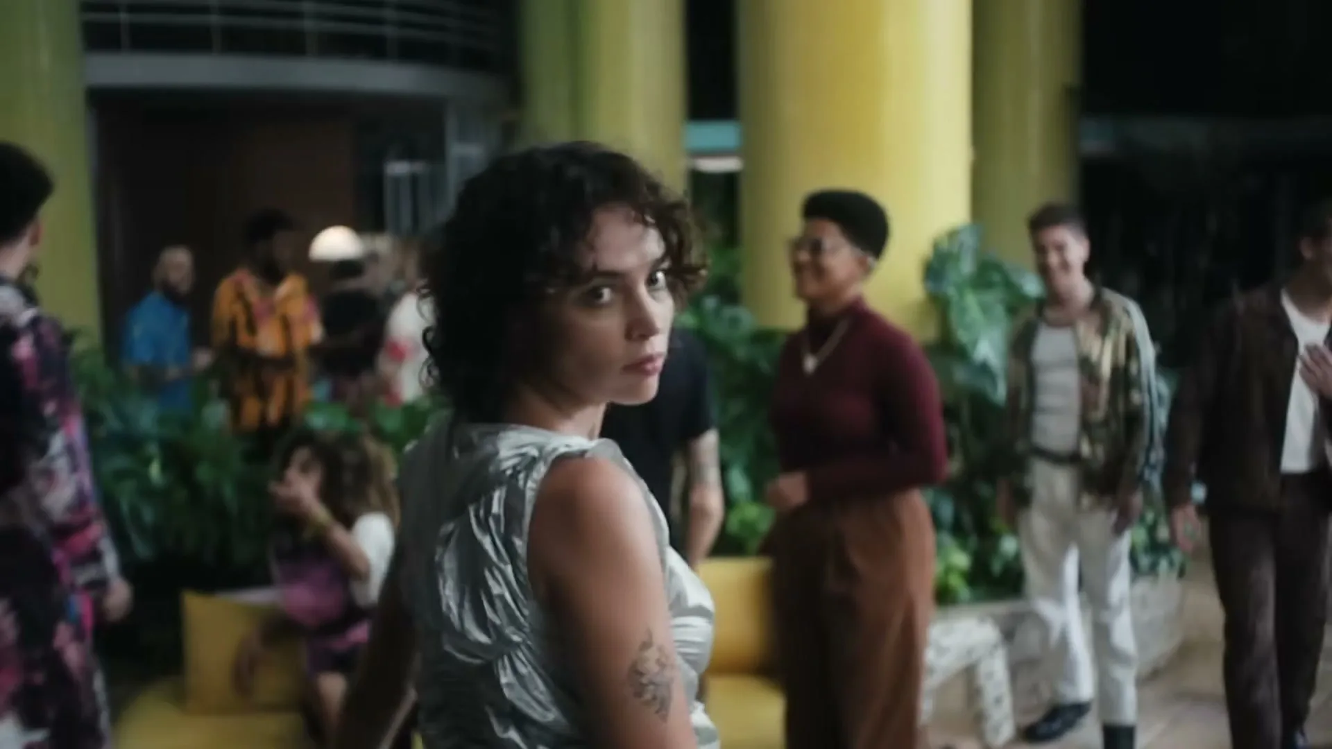 A young woman with curly hair in a silver dress walks through a crowded, vibrant hotel lobby, turning to look back at the camera.
