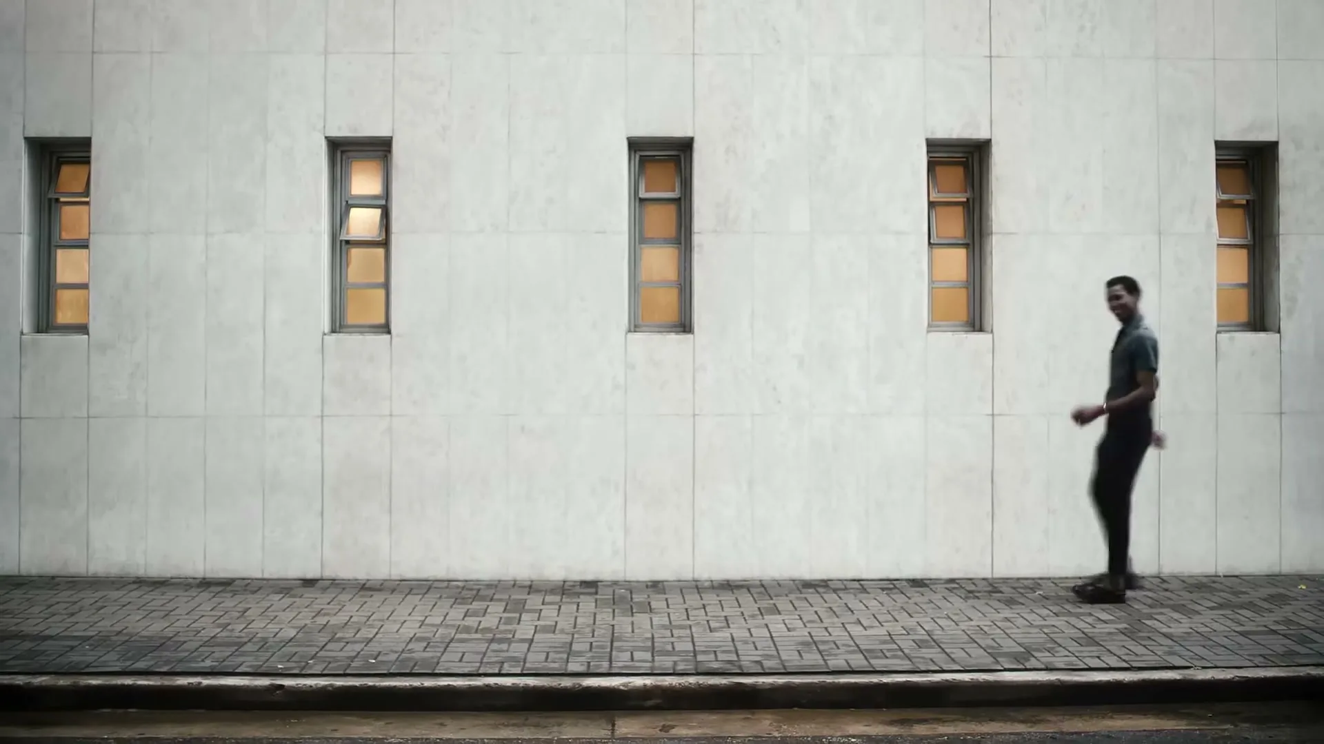 A man in dark clothing walks quickly from right to left along a sidewalk in front of a white building with several windows.