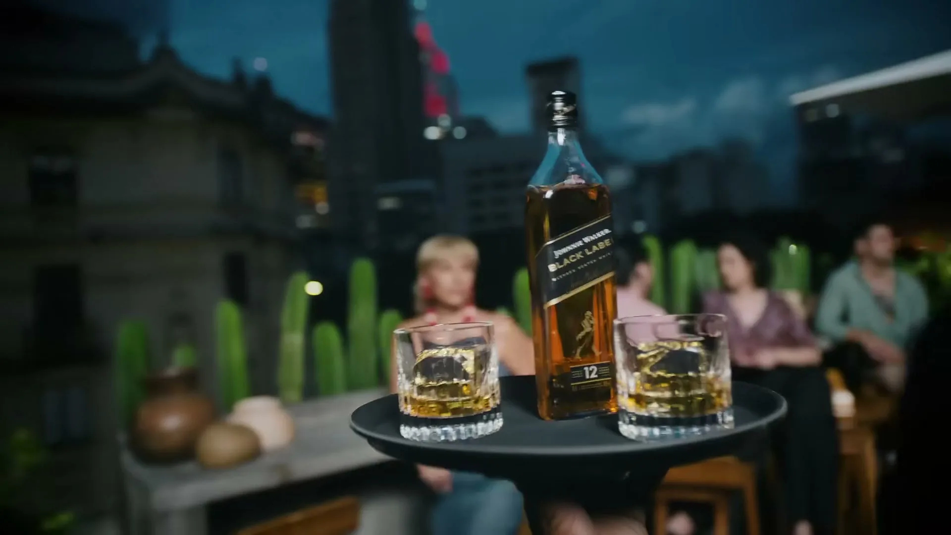 A server's hand holds a tray with a bottle of Johnnie Walker Black Label and two glasses of whiskey on a rooftop bar at dusk, as a woman in a red and white beaded necklace looks on.