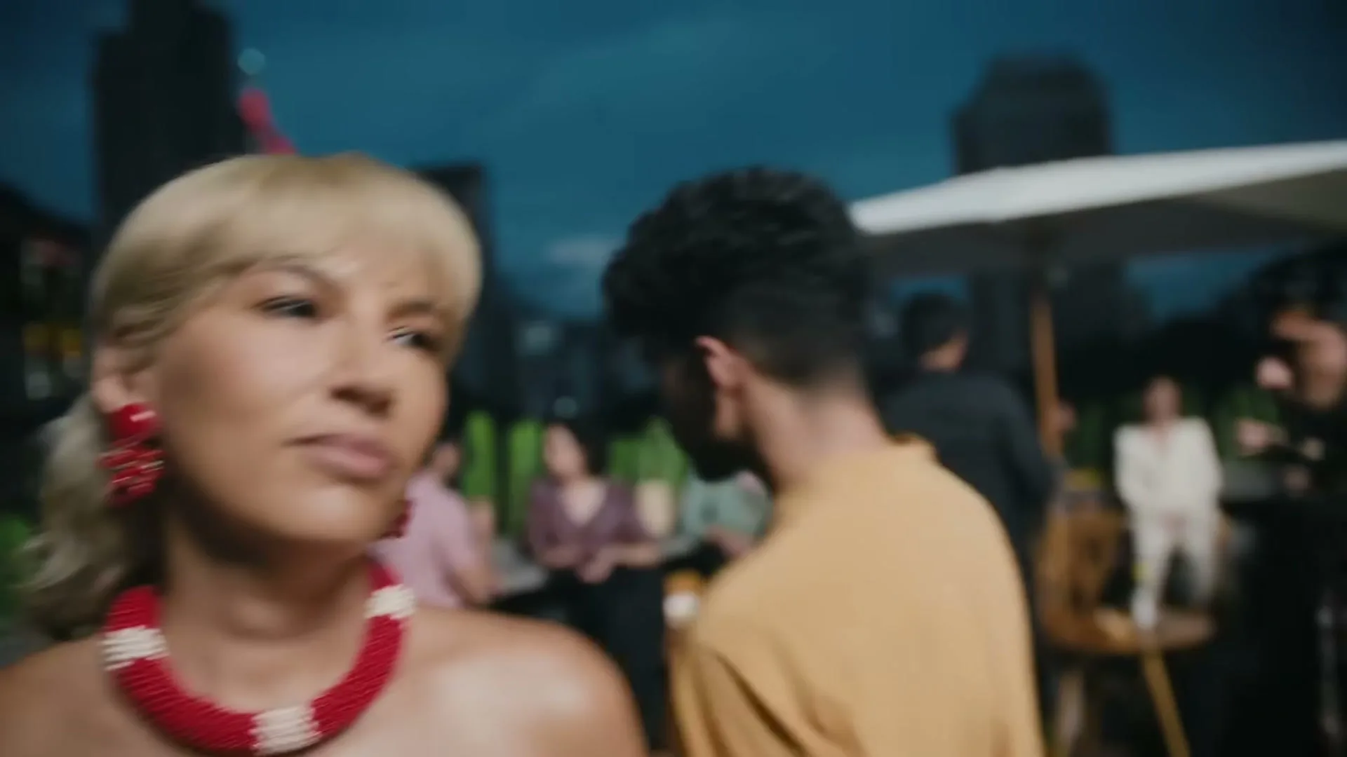 A woman with blonde hair and a red and white beaded necklace looks to the side at a rooftop party at night.