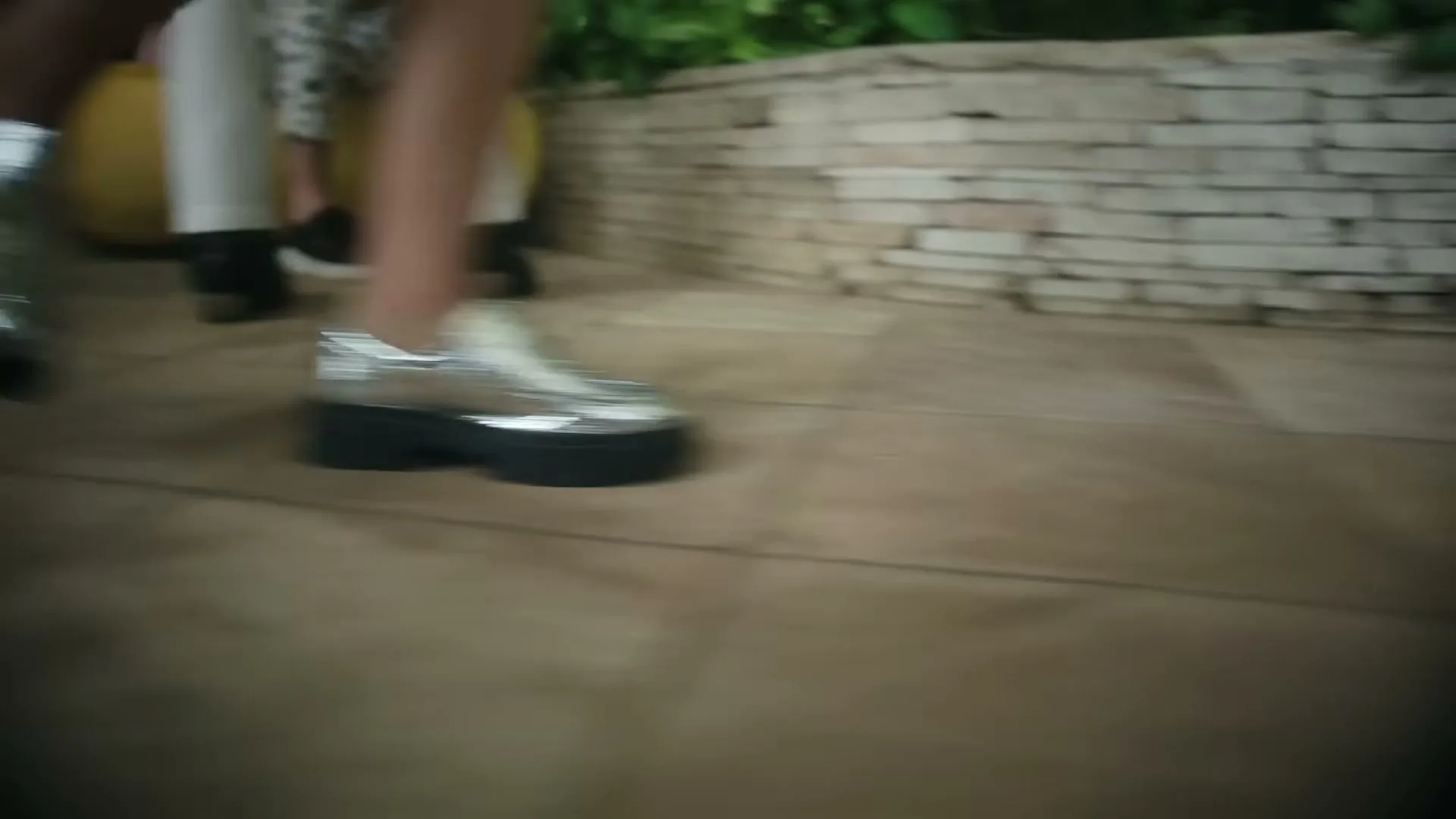 A person wearing shiny silver shoes with thick black soles walks across a tiled patio, with other people's legs visible in the background.