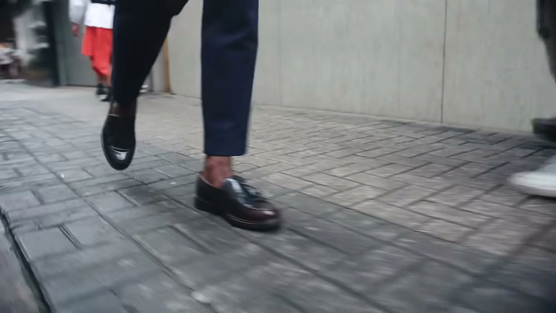 A low-angle shot of a person's legs and feet walking on a paved sidewalk, with other people's legs visible in the background.