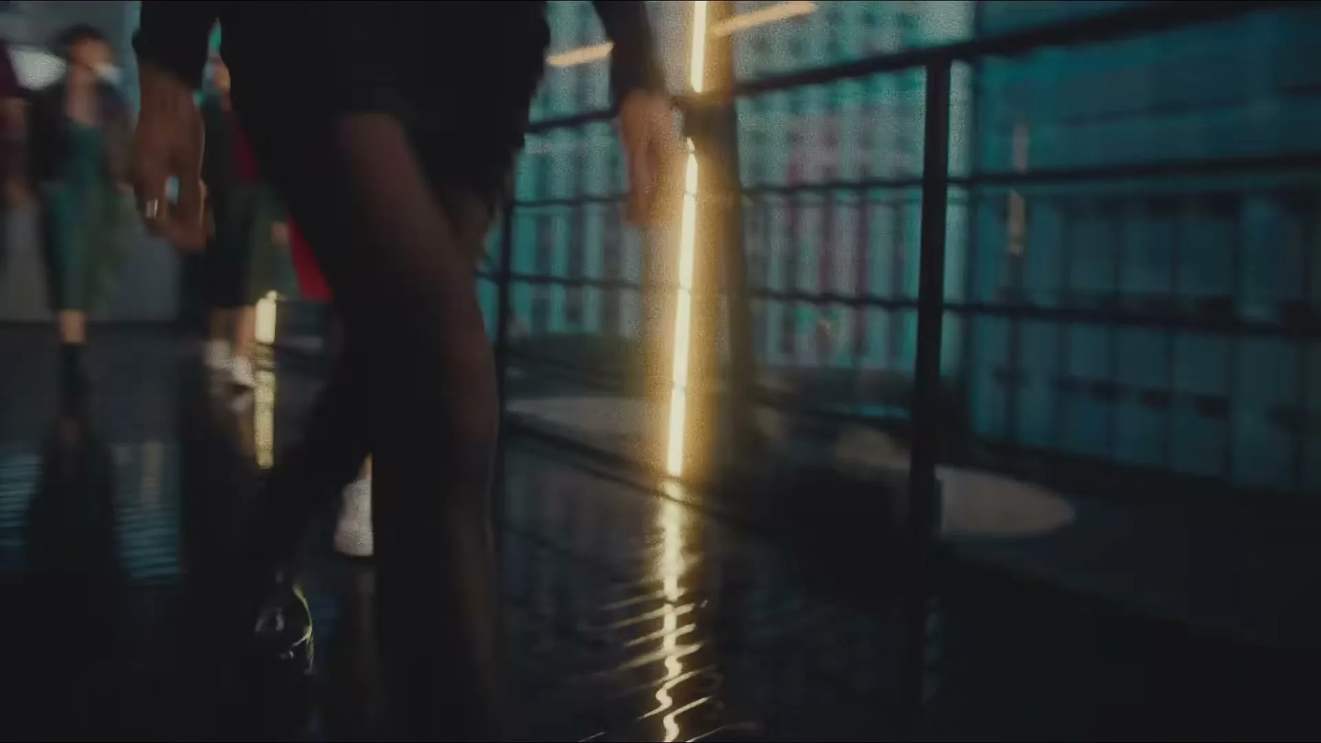A group of people walk quickly across a dark, wet floor with reflections, past a glowing vertical light and a blurred city background at night.