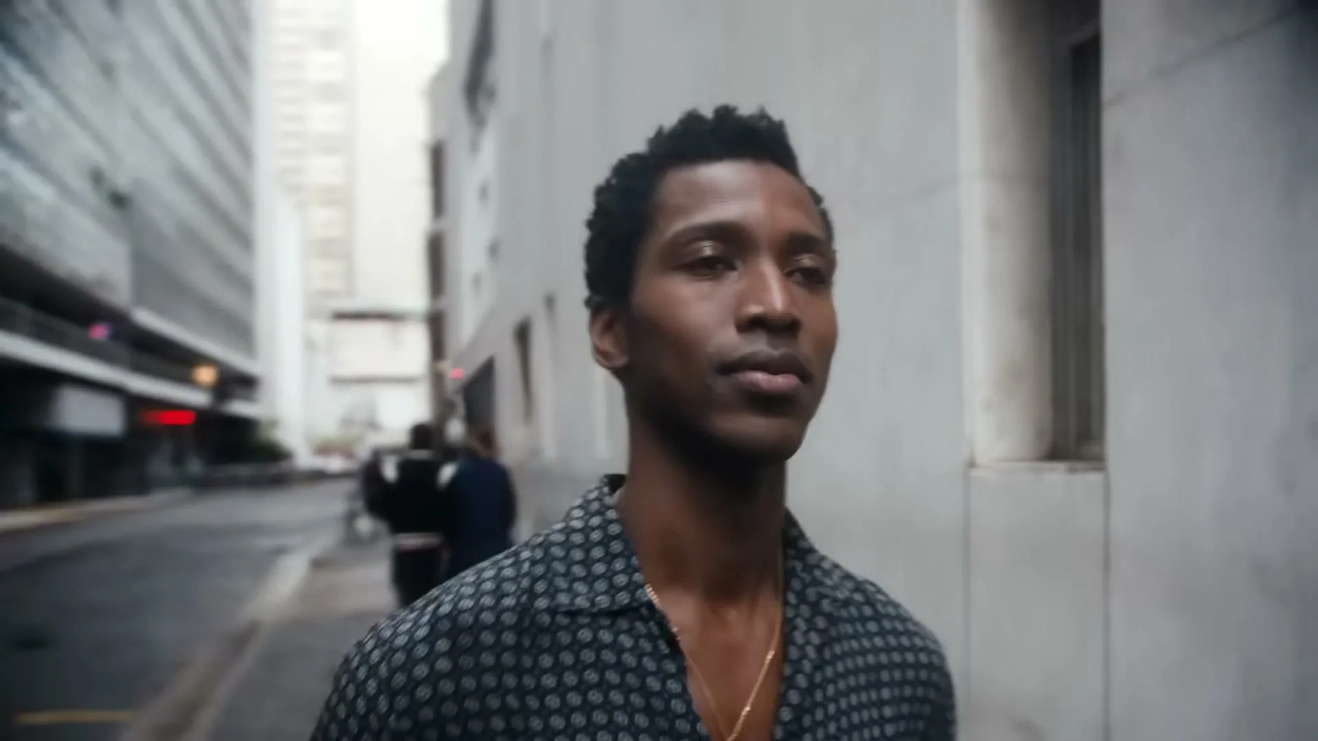 A young Black man in a patterned shirt and gold chain walks down a city street, looking forward with a determined expression.