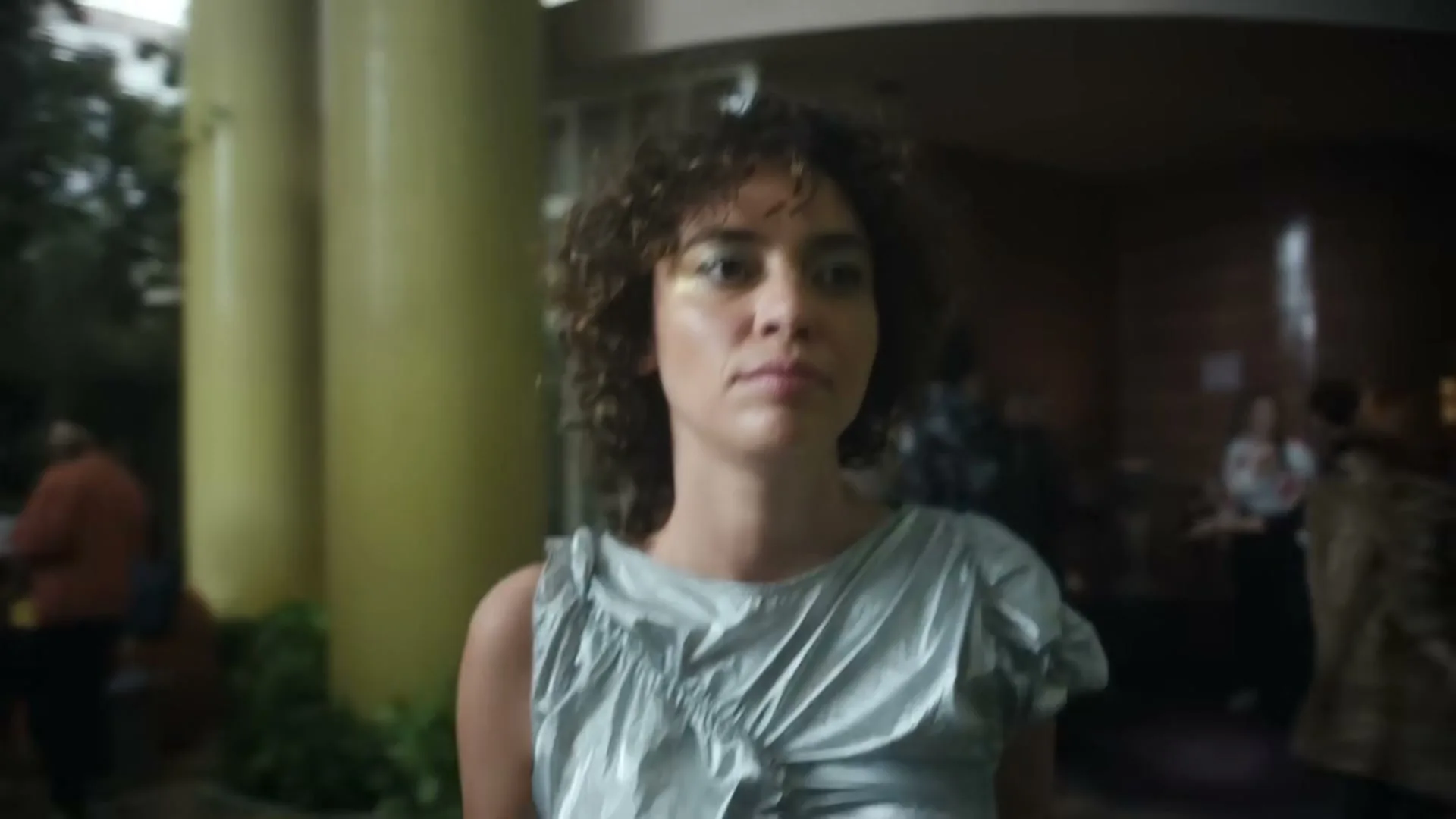 A young woman with curly hair and a silver dress walks forward, looking determined, as people mill about in the background of a building entrance.