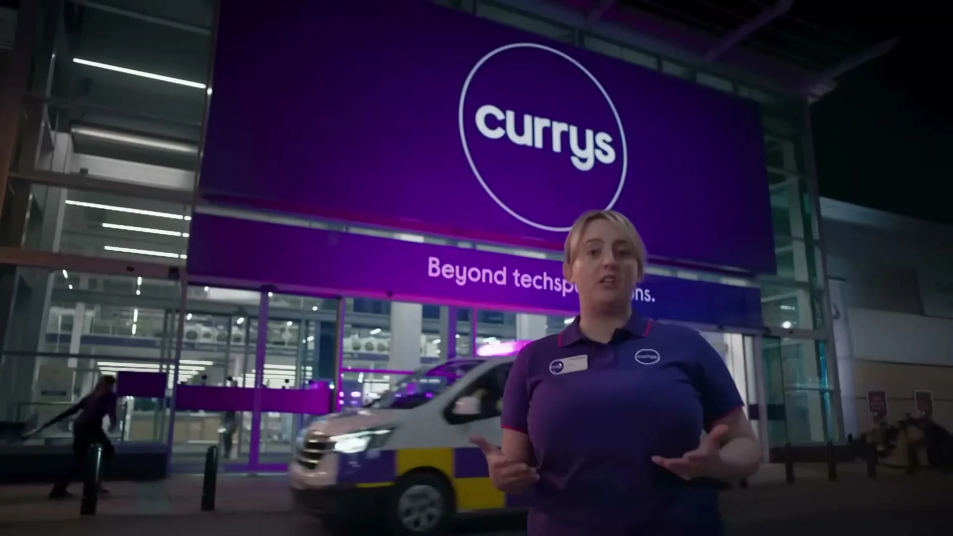 A female Currys employee in a purple uniform stands outside a Currys store at night, talking to the camera as a Currys delivery van drives past.