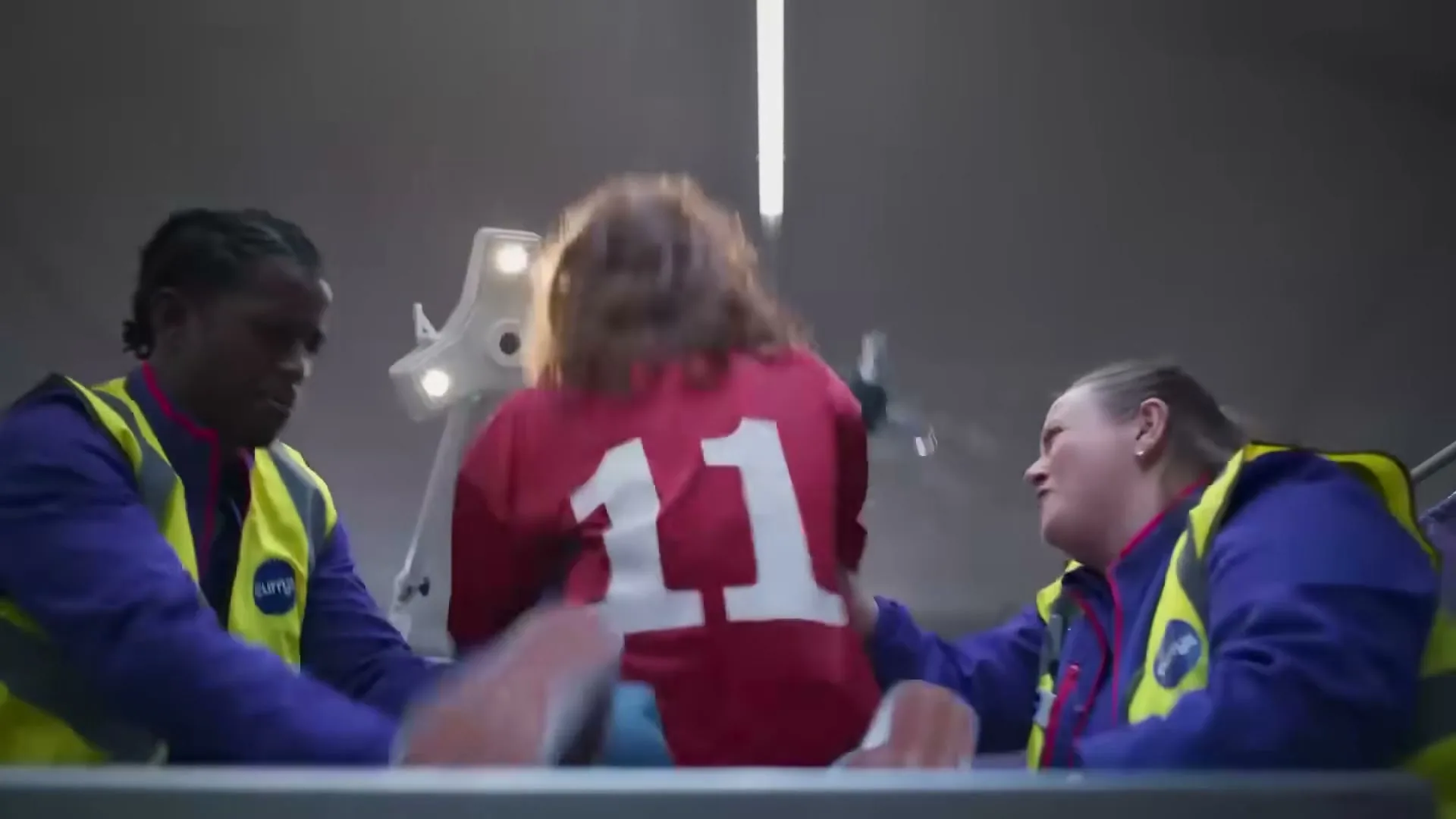 Two Currys employees in uniform help a person in a red jersey with the number 11 onto a moving platform in a large, brightly lit warehouse.