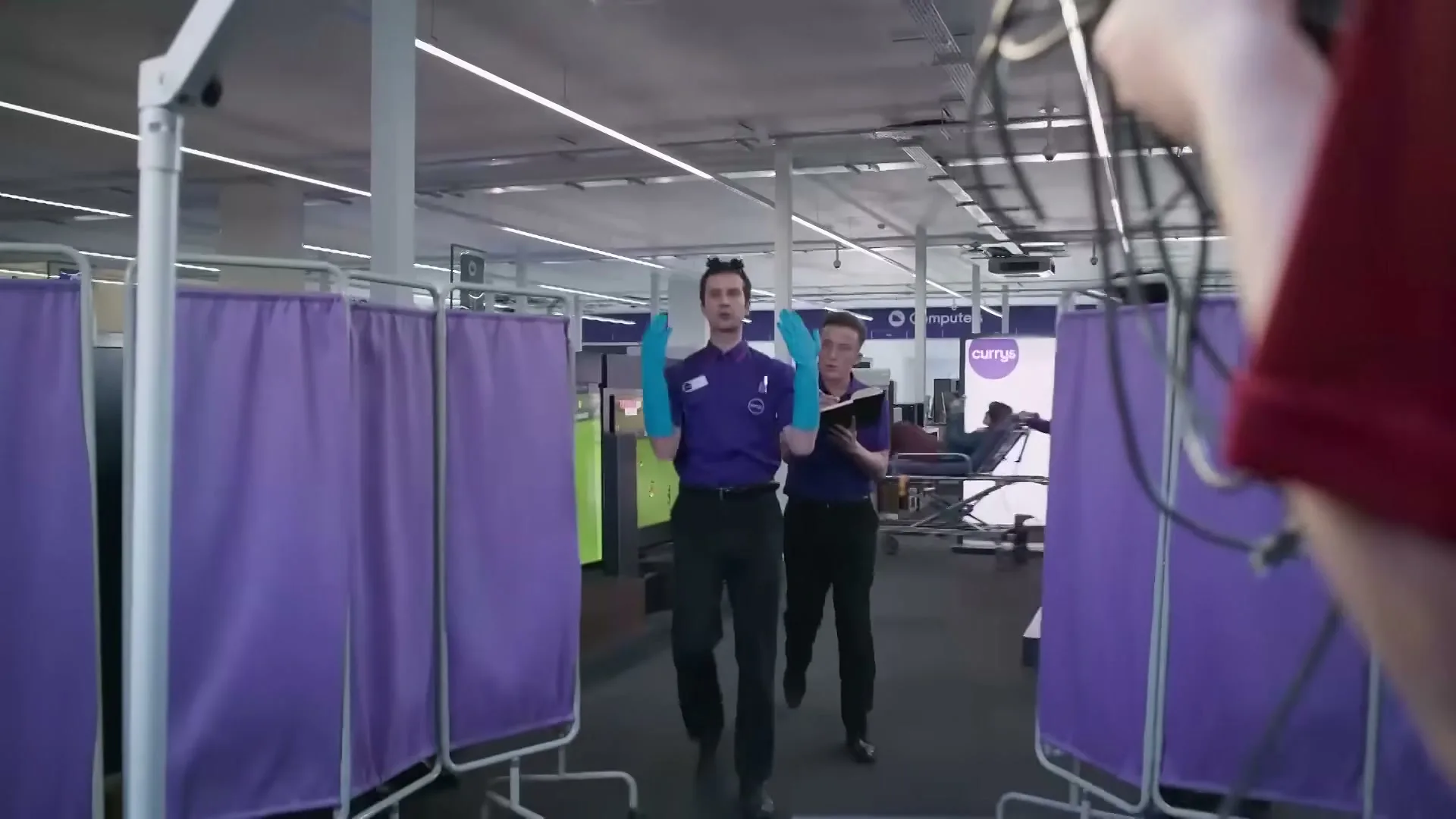 Two male employees in purple uniforms and blue gloves walk towards the camera in a tech store, one holding a clipboard.