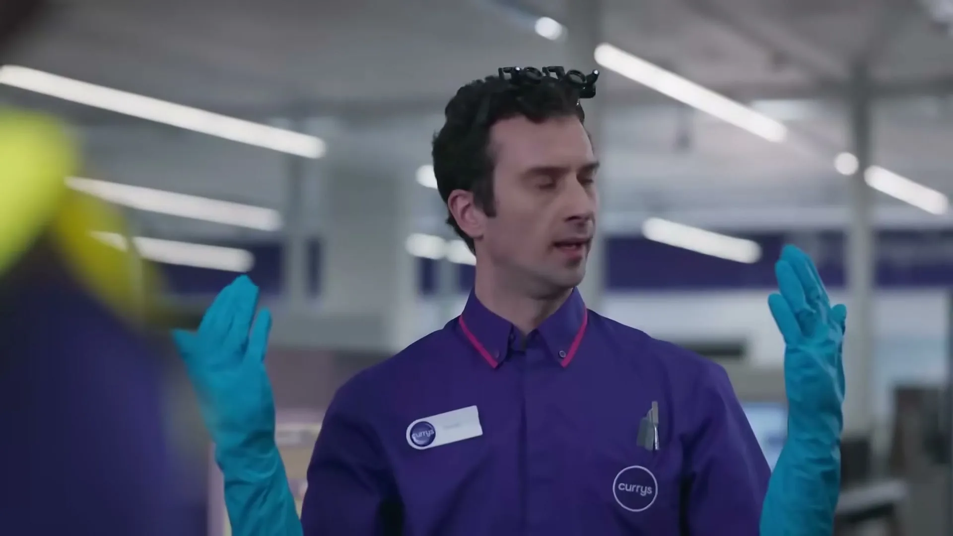 A male Currys employee in a purple uniform and blue gloves talks to a customer in a brightly lit electronics store.