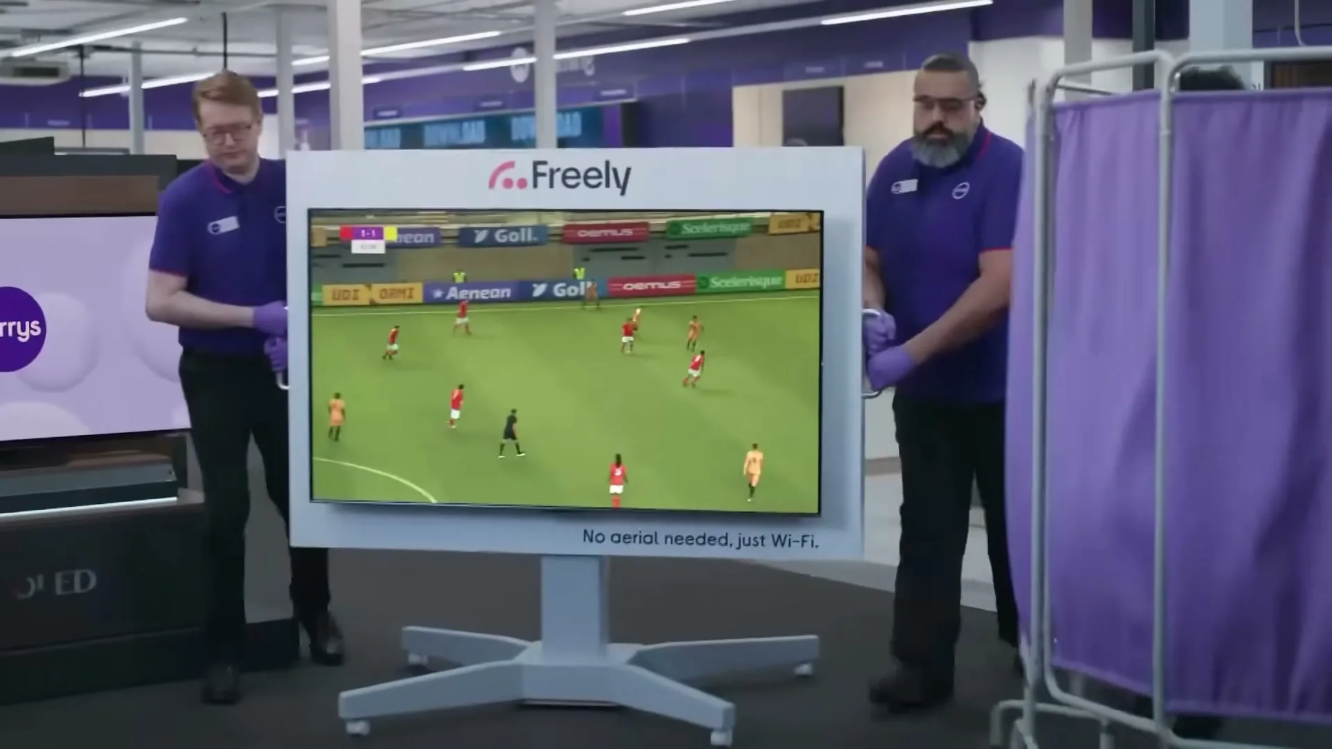 Two male Currys employees in purple shirts and black trousers move a large TV displaying a football match in a brightly lit electronics store.