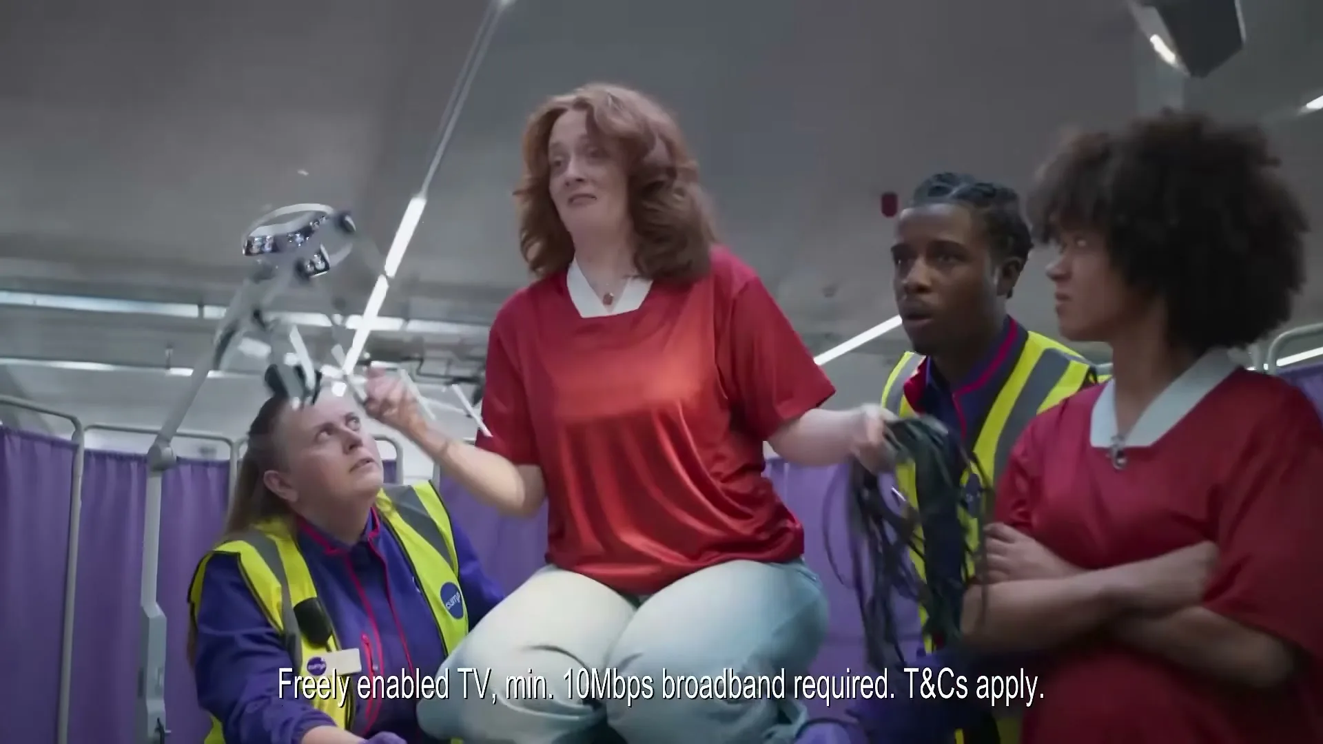 A woman with red hair in a red shirt and light blue pants sits on a table, holding an antenna and wires, surrounded by three people in yellow vests in a room with purple curtains.