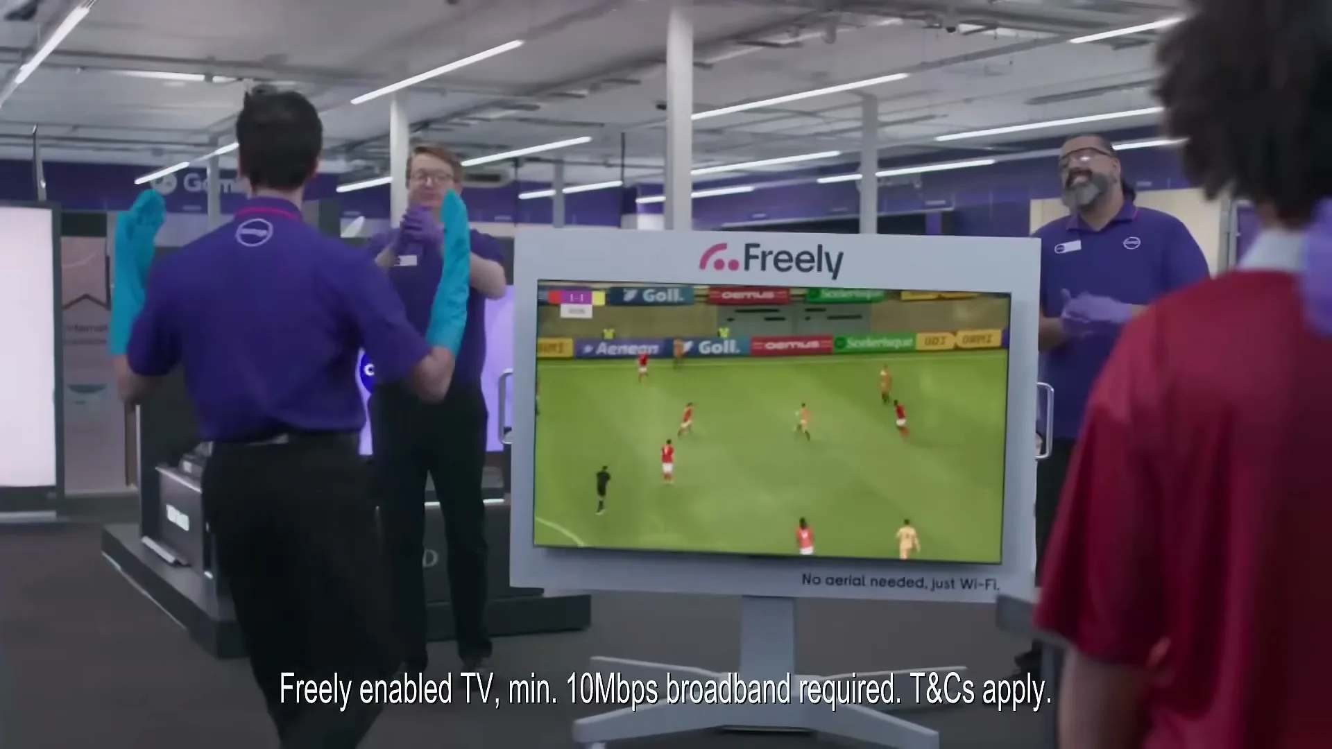 Three male store employees in purple shirts and blue gloves dance around a TV displaying a soccer game in an electronics store.