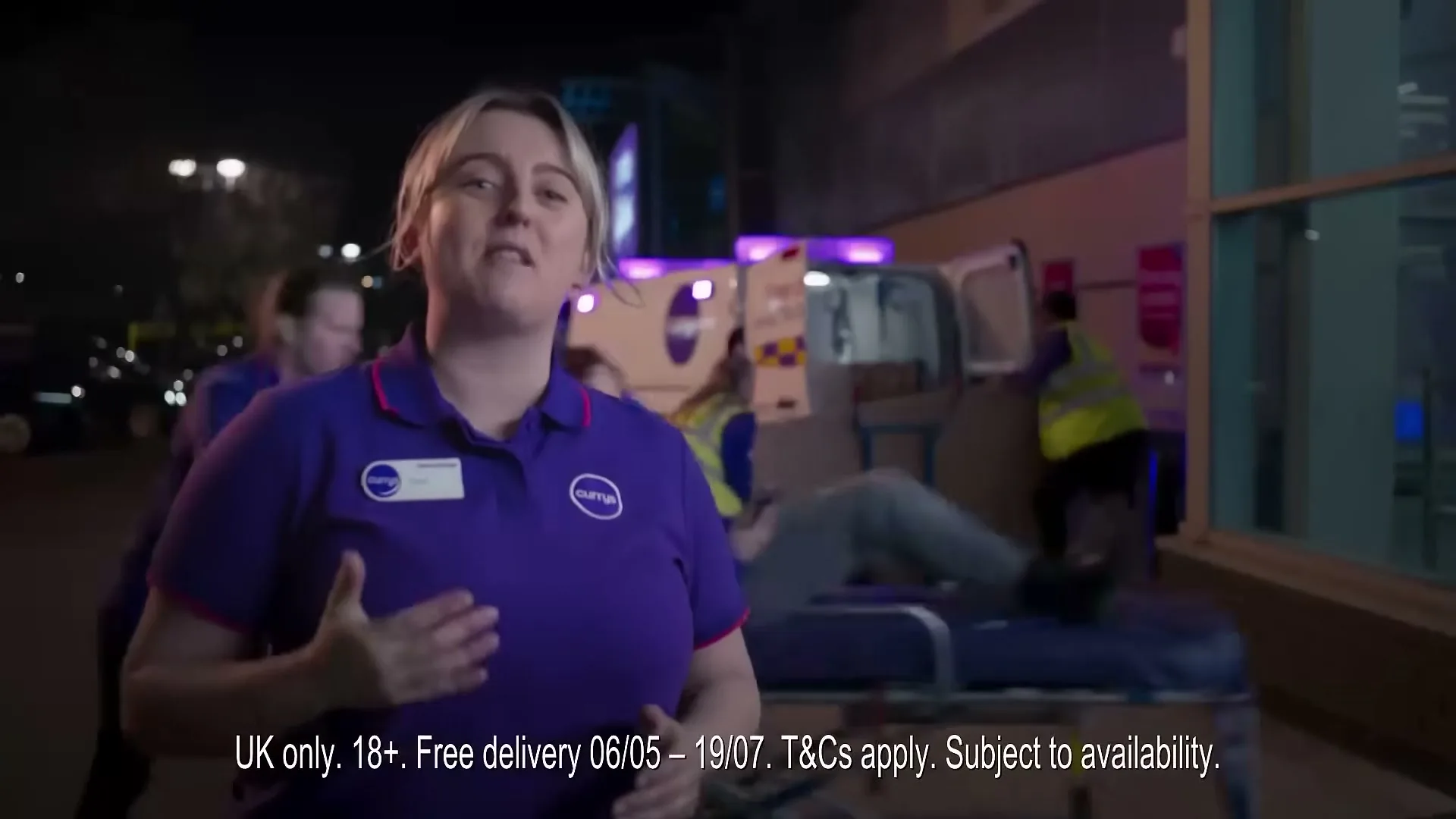 A female Currys employee in a purple uniform speaks directly to the camera, while other employees unload a delivery van in the background at night.