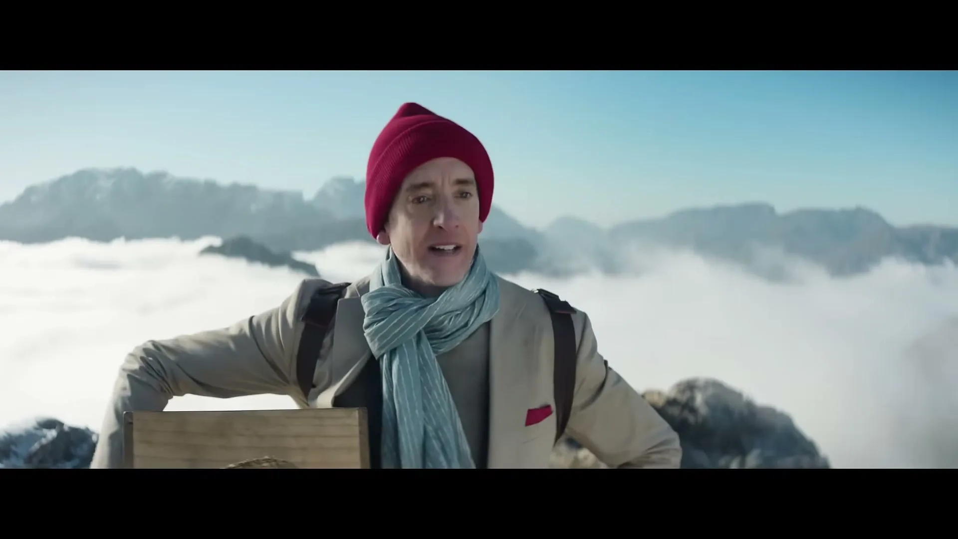 A man in a red beanie and scarf stands on a snowy mountain peak, looking out over clouds and distant mountains.