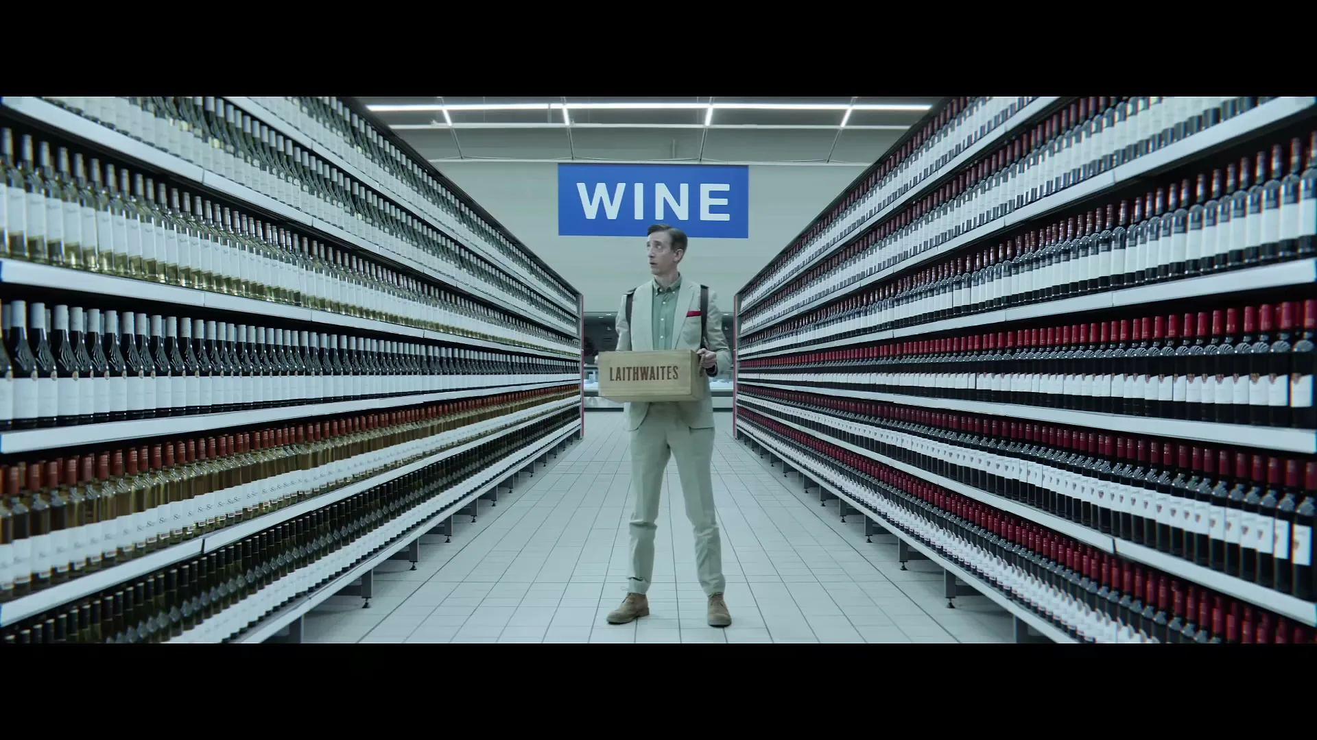 A man in a beige suit stands in a supermarket wine aisle, holding a box of Laithwaites wine, looking up at the "WINE" sign.