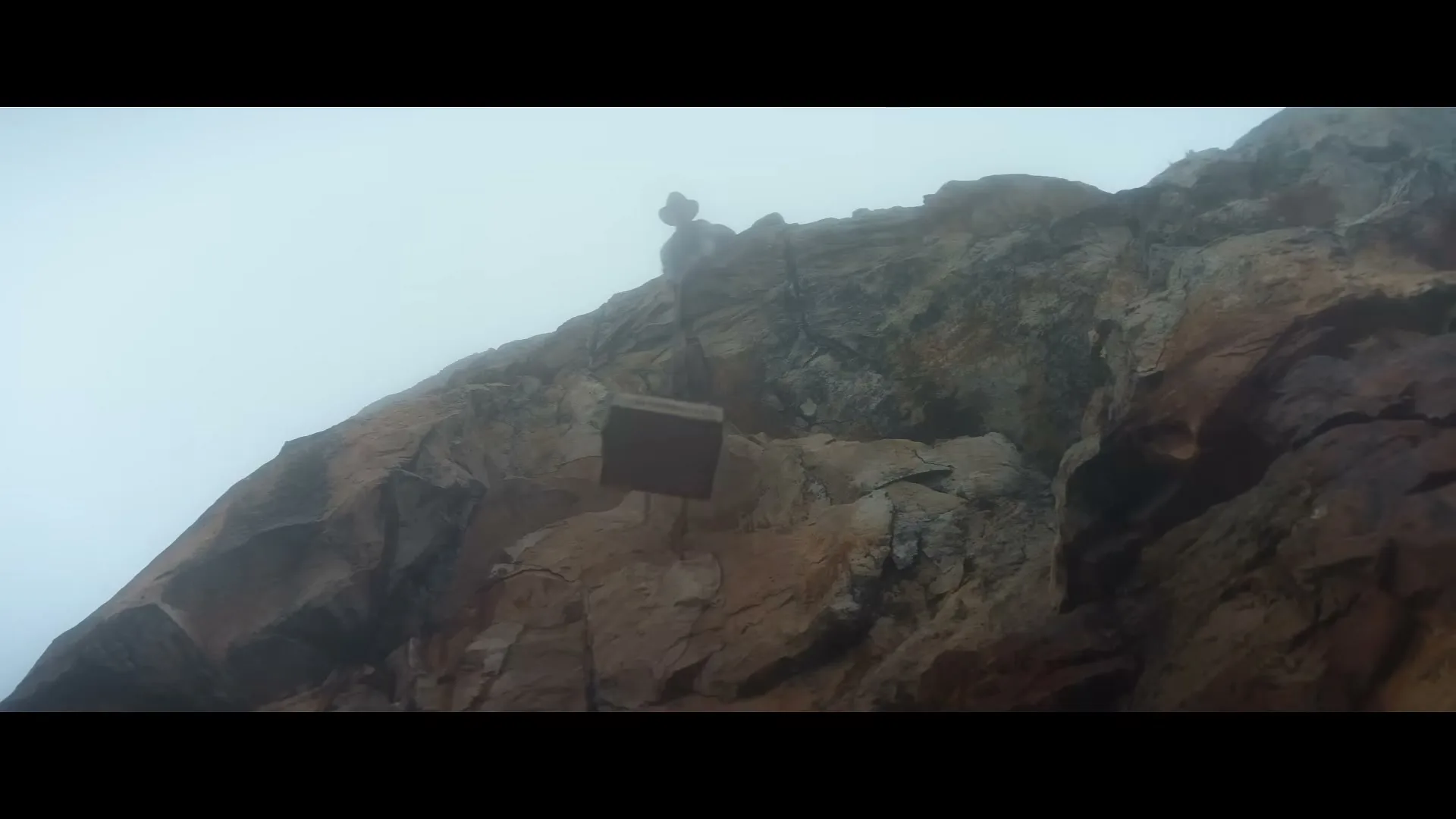 A small, square, brown sign with "LAITHWAITES" written on it is lowered down a rocky cliff face on a foggy day.