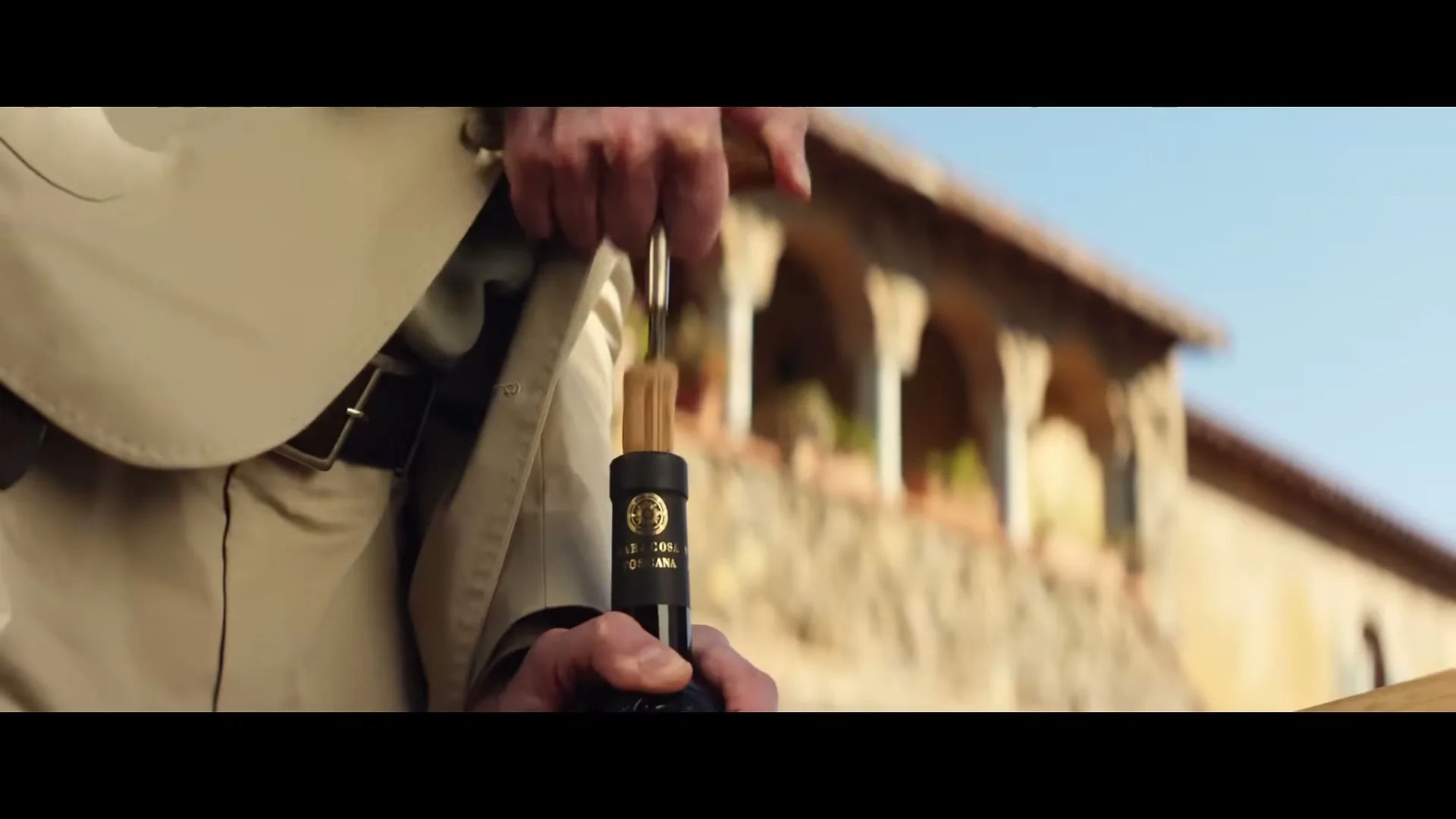 A man in a beige trench coat uses a corkscrew to open a bottle of red wine, with a stone building in the background.