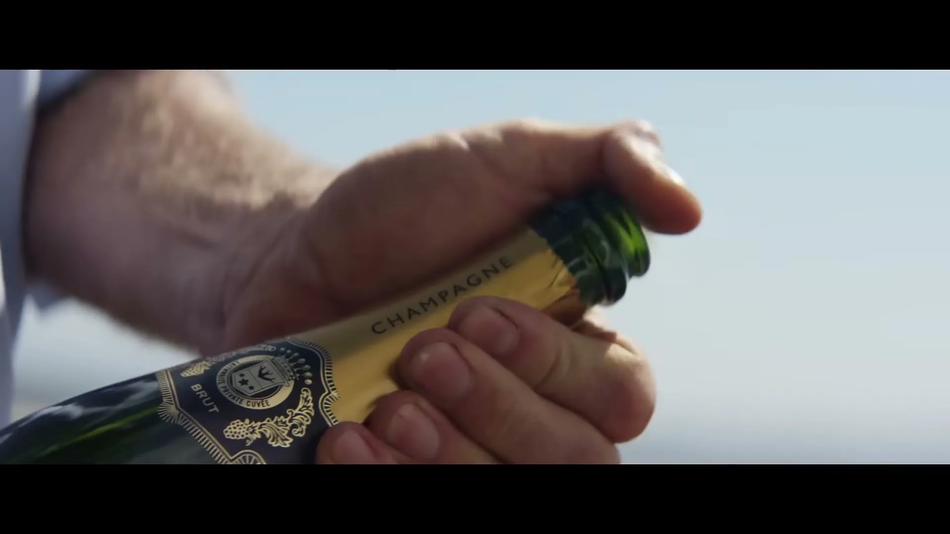 A man's hands remove the cork from a bottle of champagne outdoors on a sunny day.