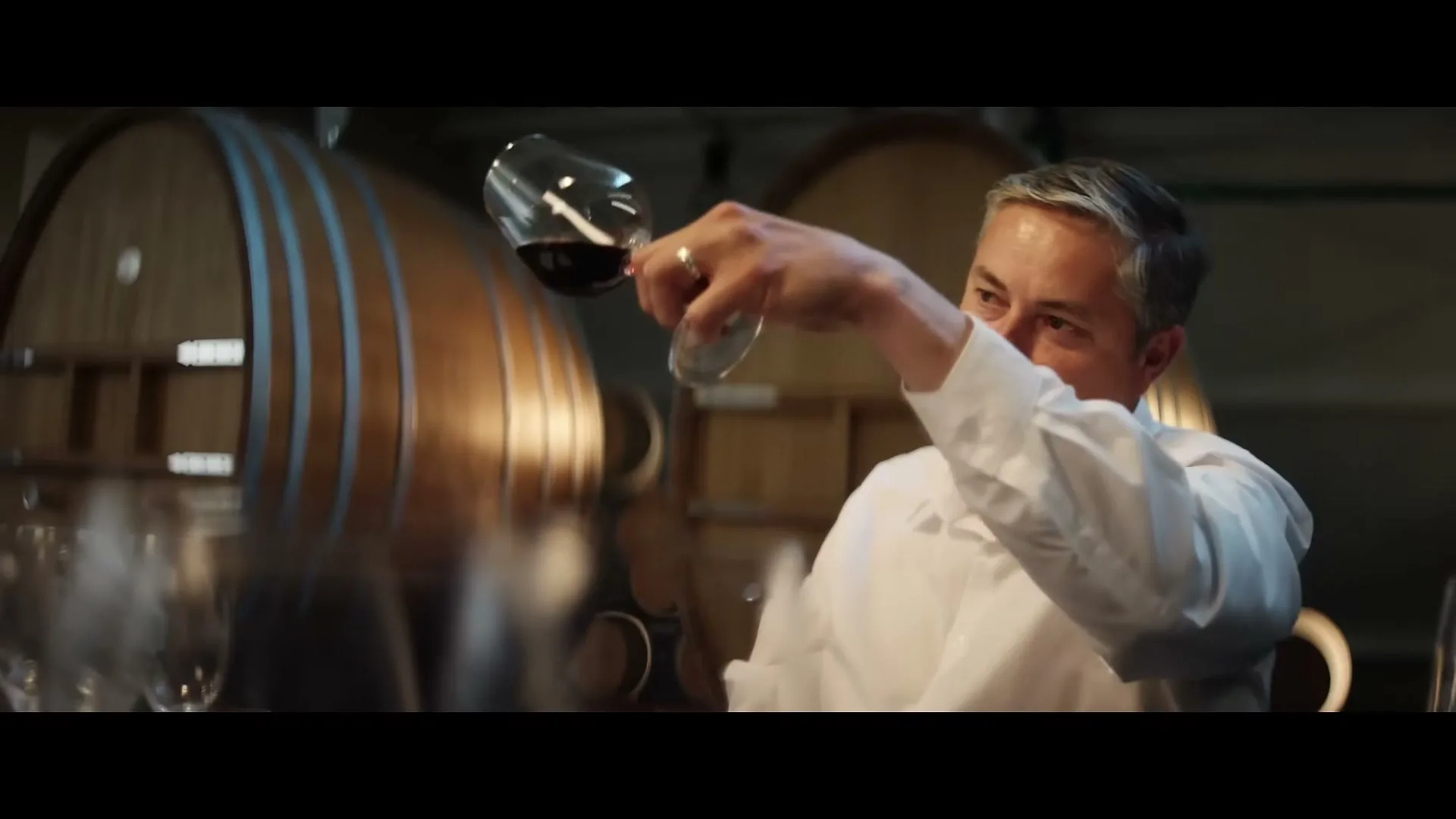 A male sommelier in a white shirt swirls red wine in a glass, inspecting it in a dimly lit wine cellar with large wooden barrels.