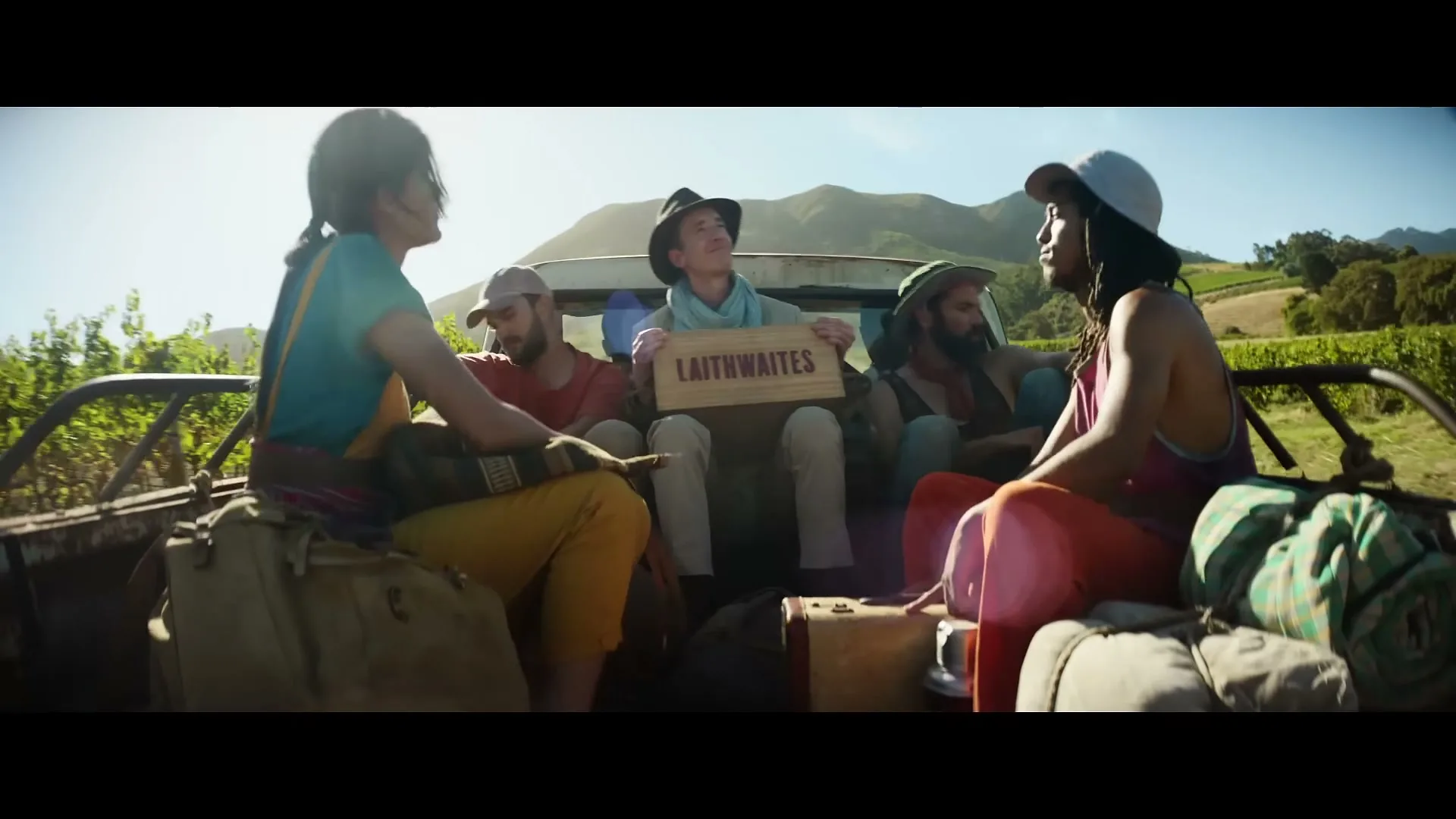 A diverse group of five friends, dressed in casual attire, ride in the back of a pickup truck through a sunny vineyard, holding a "Laithwaites" sign.