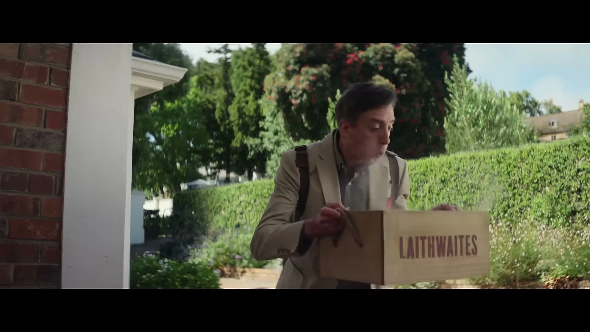 A man in a suit carries a Laithwaites box, blowing on it to remove dust, as he walks towards a house on a sunny day.