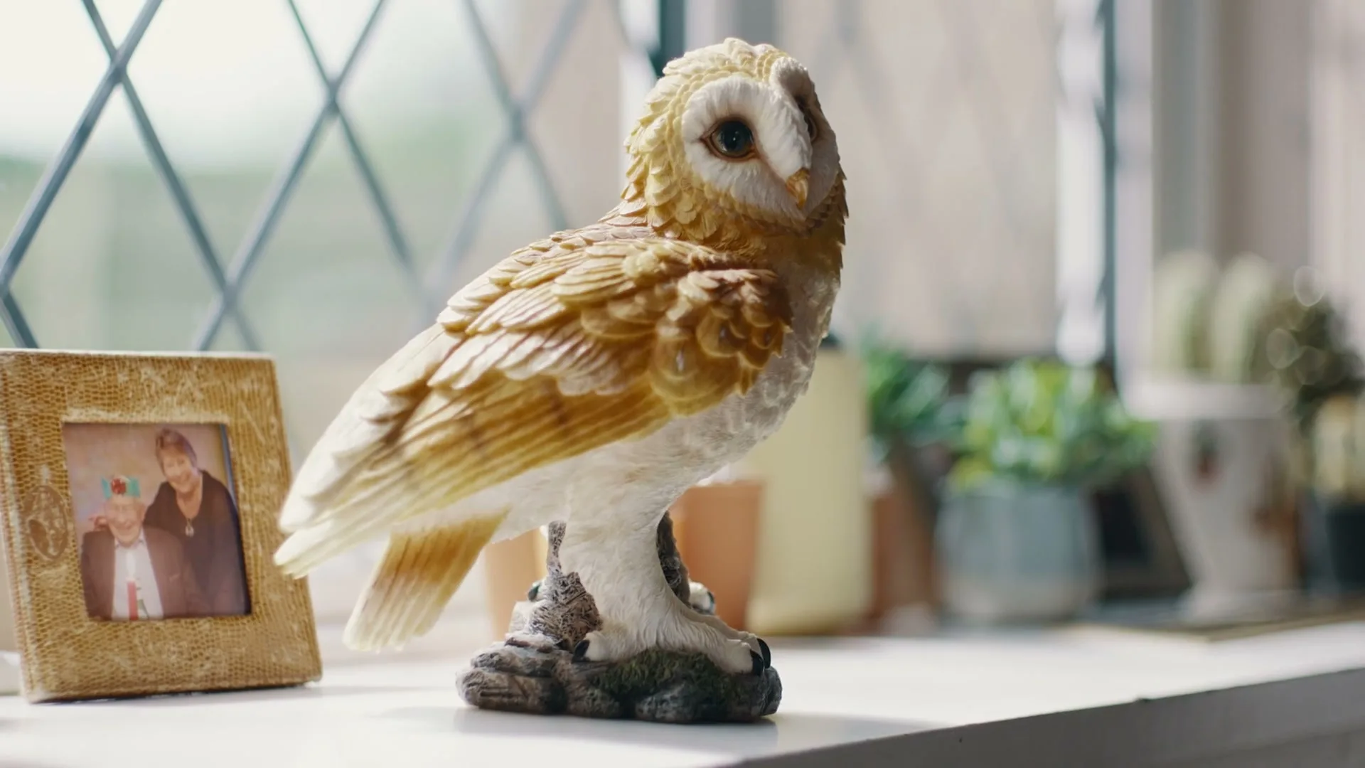 A ceramic owl figurine sits on a windowsill next to a framed photo of two people, with a leaded glass window in the background.