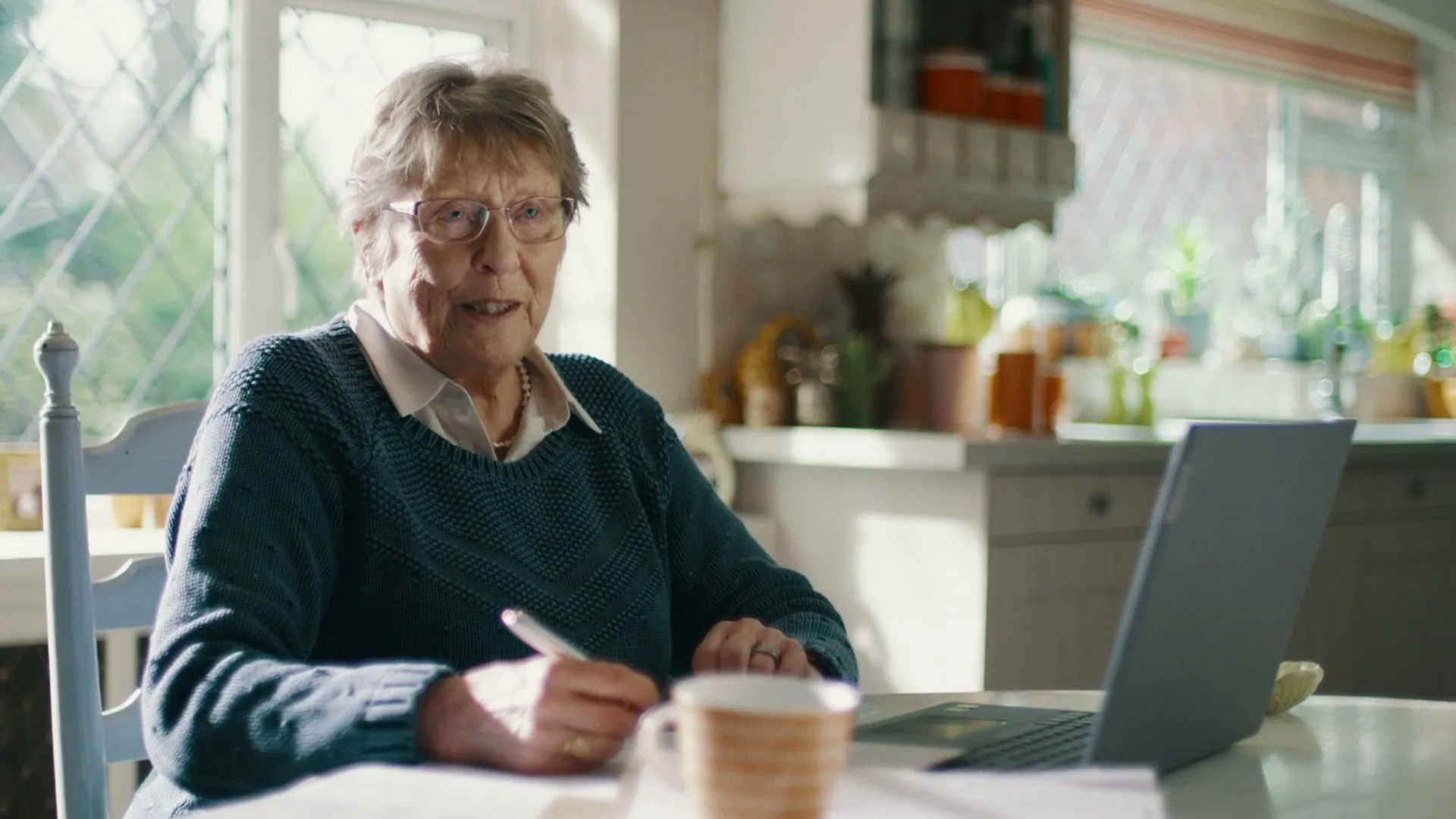 An elderly woman with glasses and a teal sweater sits at a table, writing on paper while looking at a laptop in a sunlit kitchen.