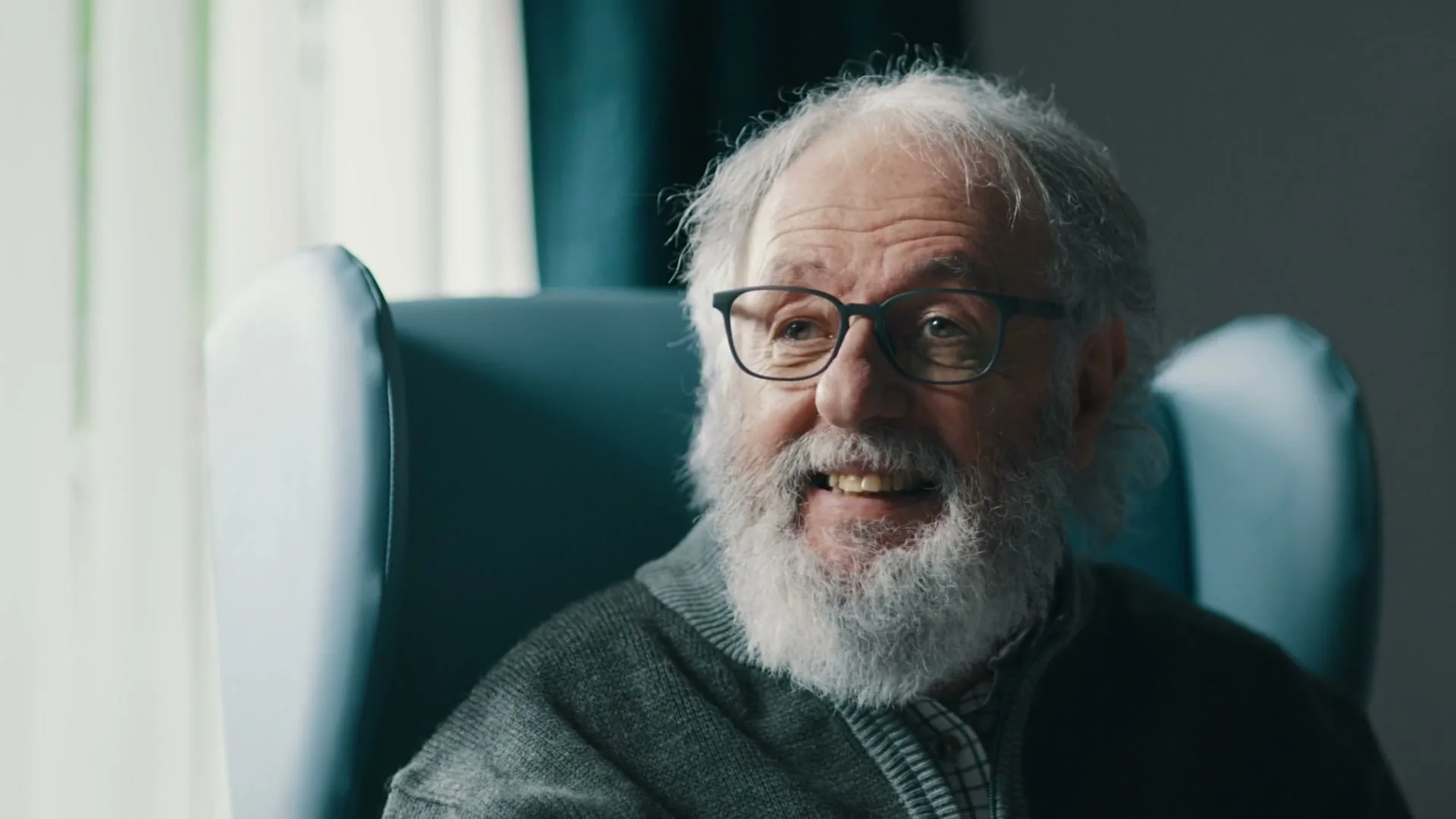 An elderly man with a white beard and glasses sits in a blue armchair, smiling and looking off-camera in an interior setting.