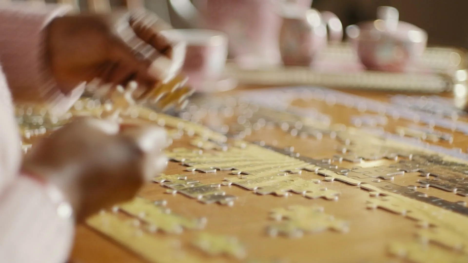 A person with dark skin and a pink sweater is seen from a medium closeup, assembling a gold jigsaw puzzle on a wooden table in an interior setting.