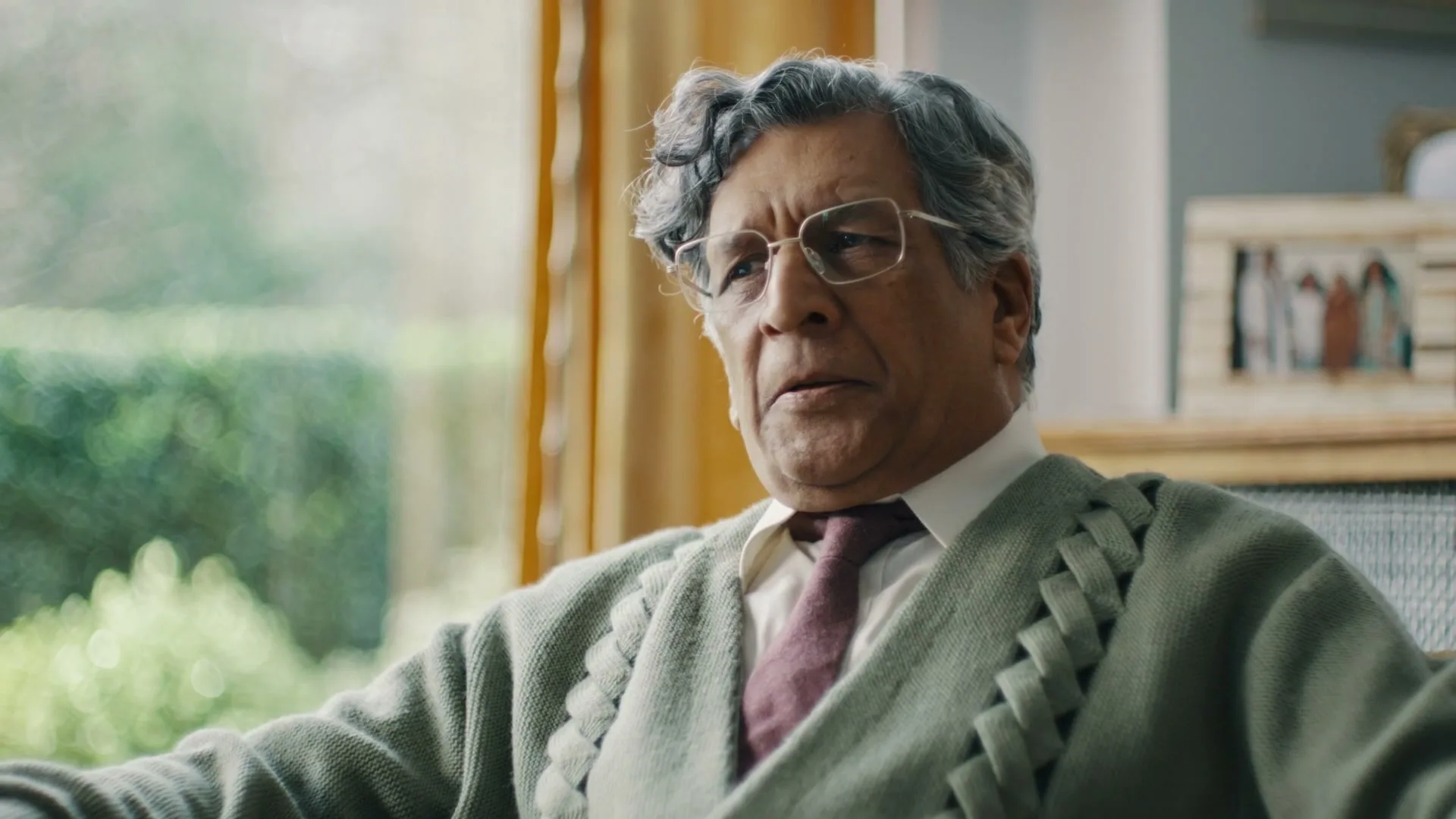 An older man with grey hair and glasses, wearing a green cardigan and tie, sits in a living room, looking thoughtful.