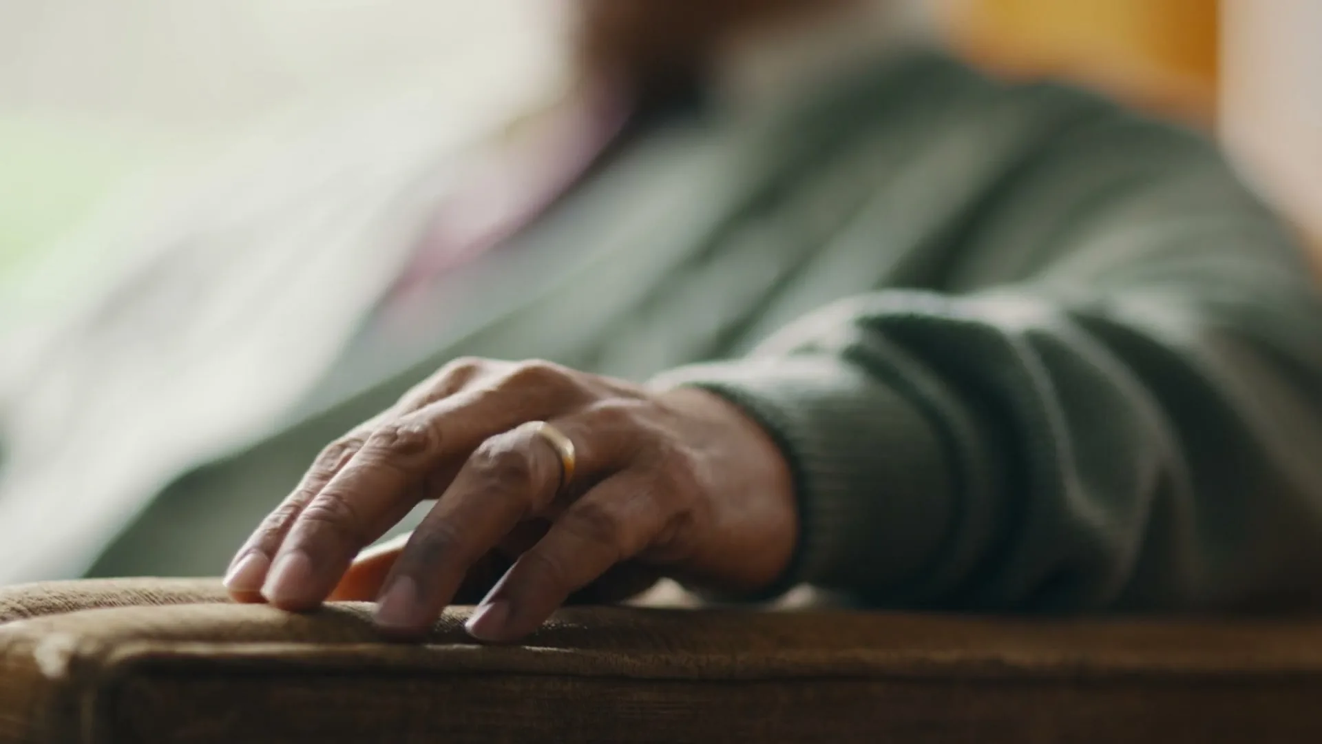 A close-up shot of an older person's hand, wearing a wedding ring, resting on a brown armchair in a softly lit interior.