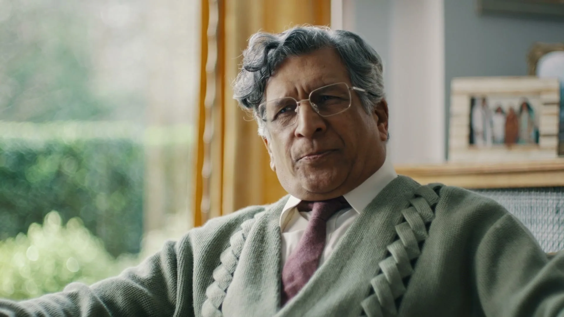 An older man with grey hair and glasses, wearing a green cardigan, sits in a living room and speaks, looking slightly off-camera.