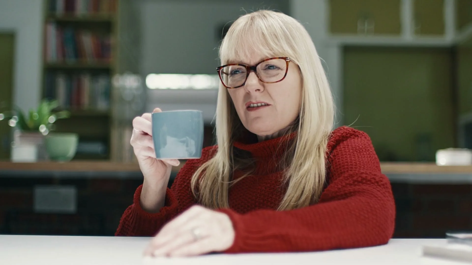 A blonde woman in her 40s, wearing a red sweater and glasses, sits at a table in a kitchen, holding a blue mug and talking.