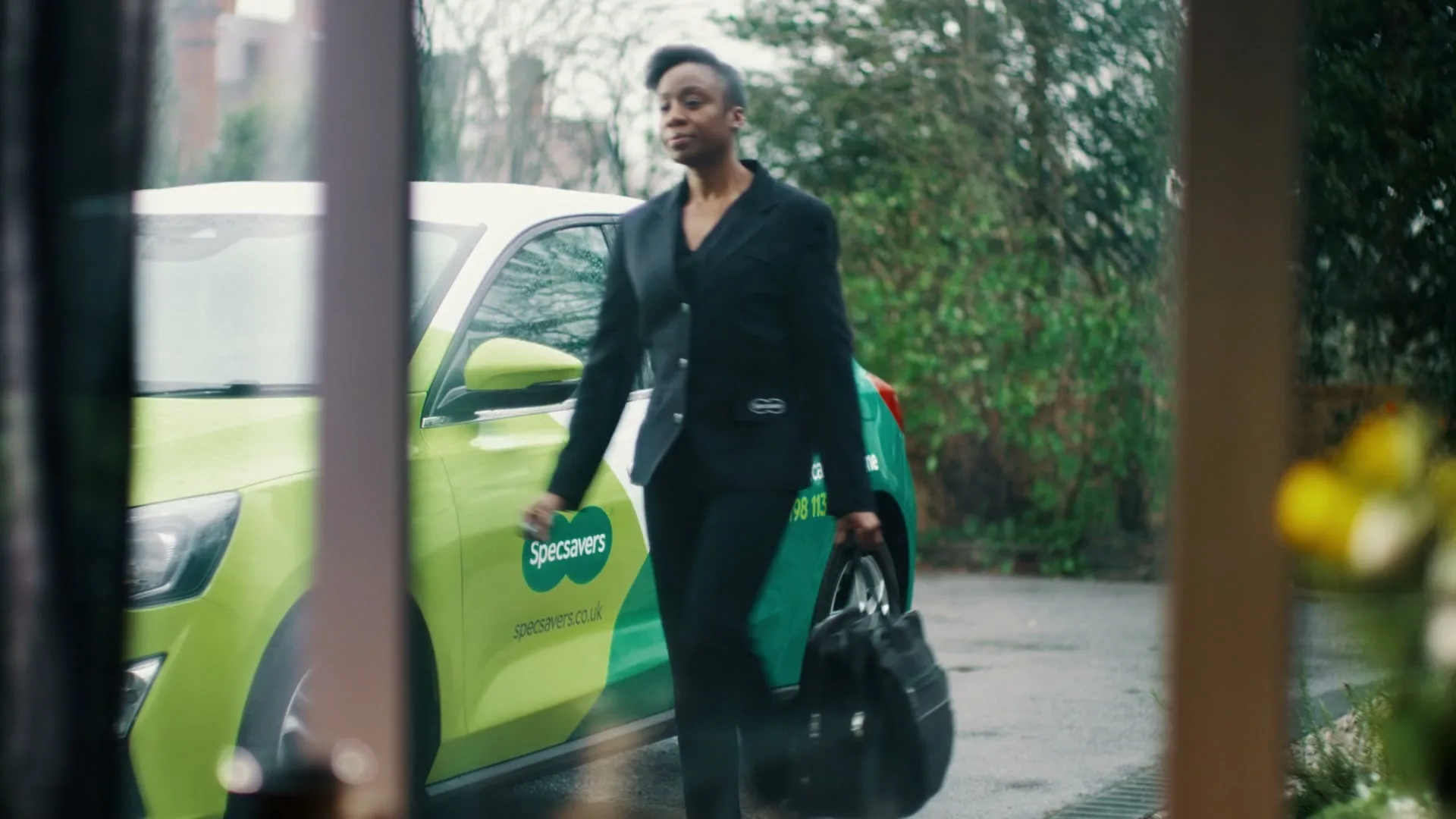 A woman in a black suit walks away from a Specsavers branded car, carrying a black bag, seen through a window on a rainy day.