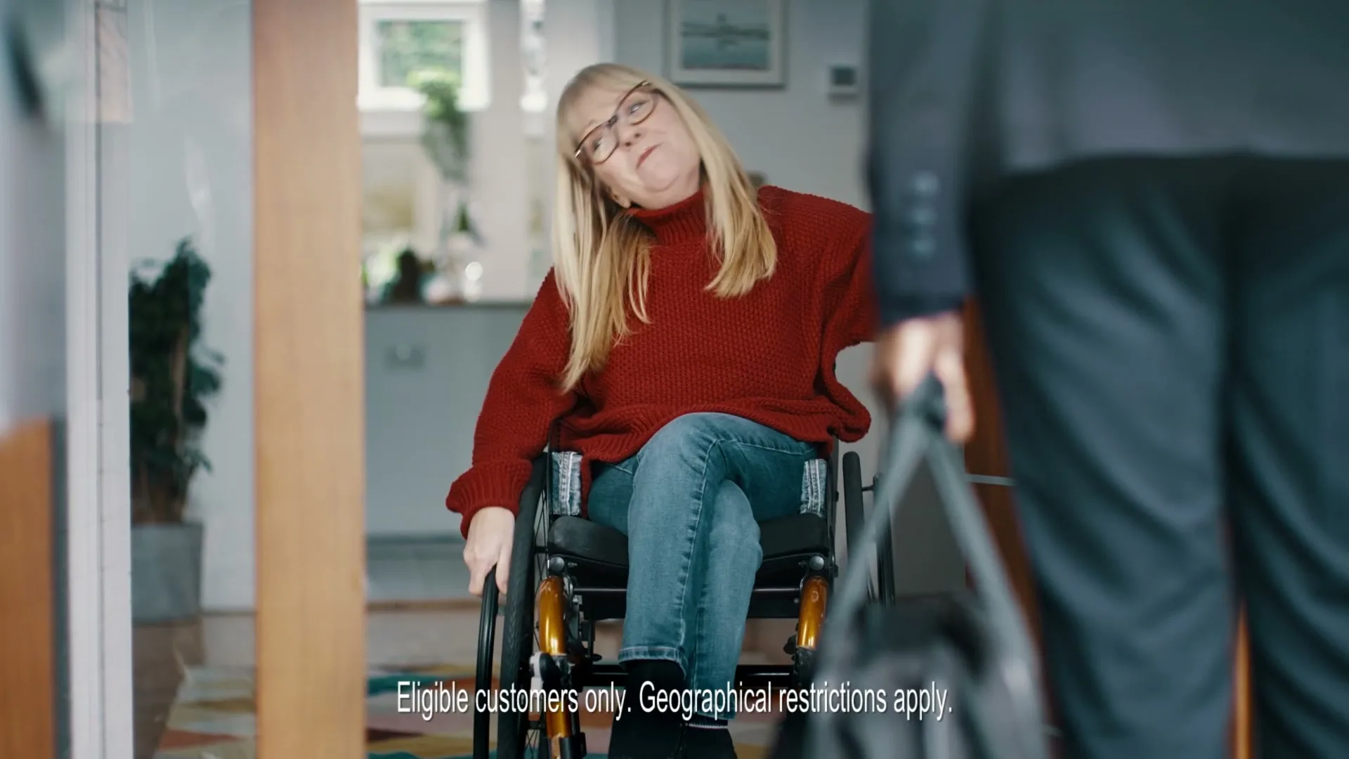 A blonde woman in a wheelchair, wearing glasses and a red sweater, smiles as she greets someone at the door of her home.