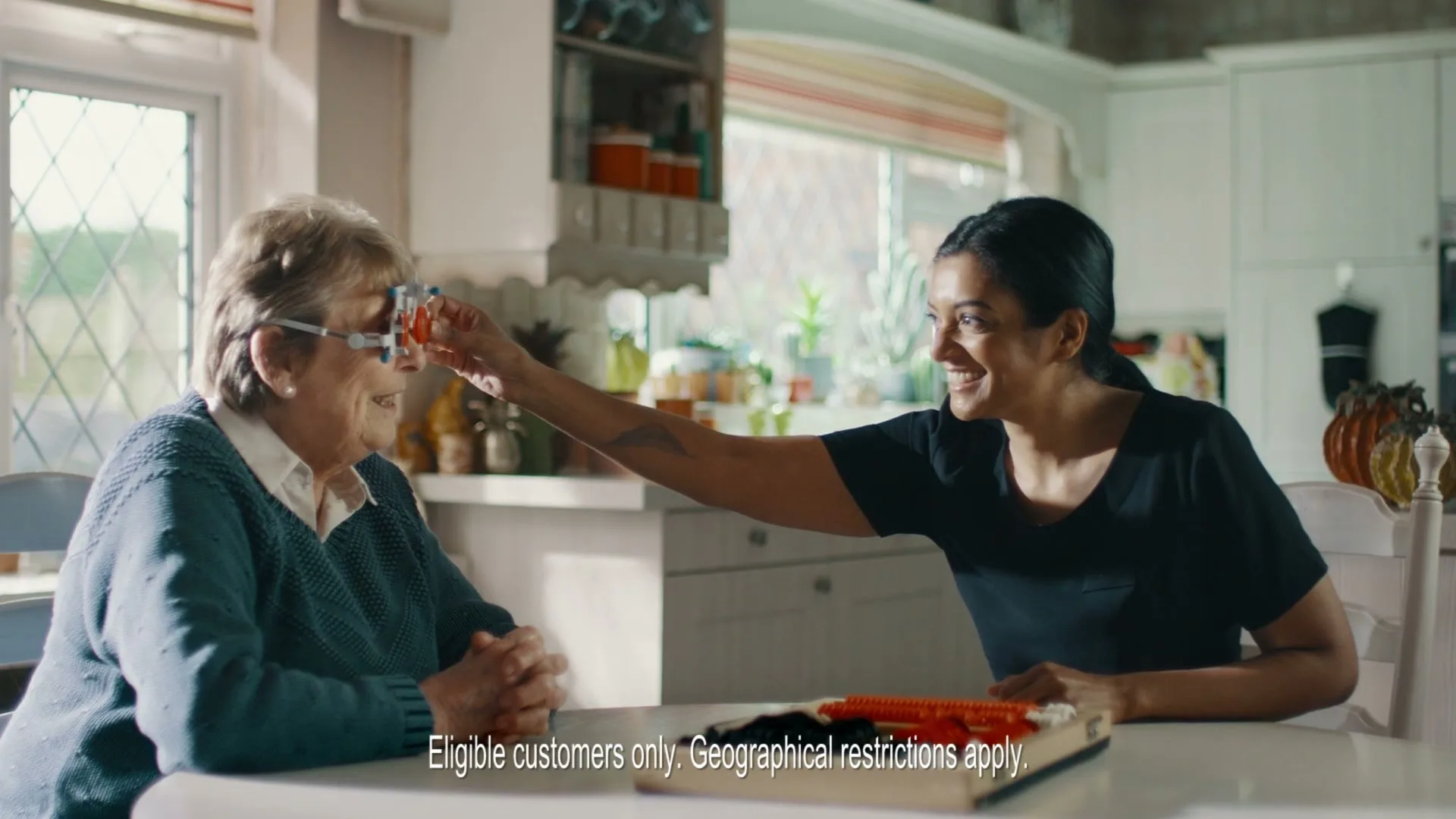 An elderly woman sits at a kitchen table while a younger woman helps her try on a pair of glasses, both smiling.