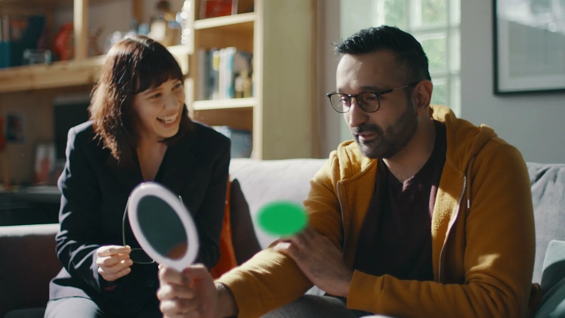 A man in glasses holds a mirror while a woman smiles at him on a sofa in a sunlit living room.
