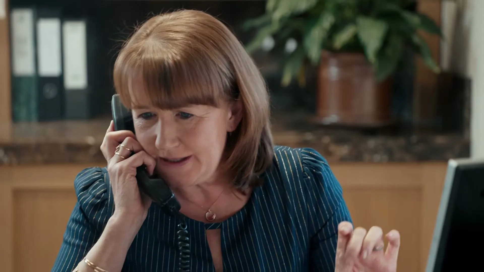 A middle-aged woman with auburn hair, wearing a blue striped blouse, talks on a landline phone at an office desk.
