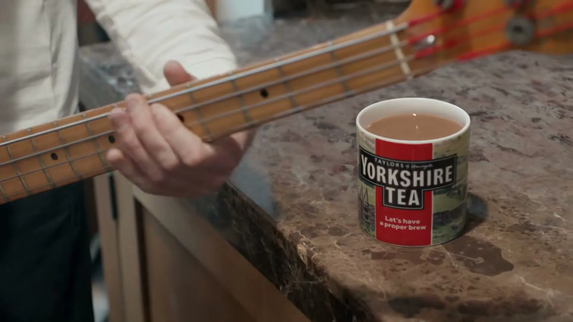 A person's hands play a bass guitar next to a mug of Yorkshire Tea on a kitchen counter.