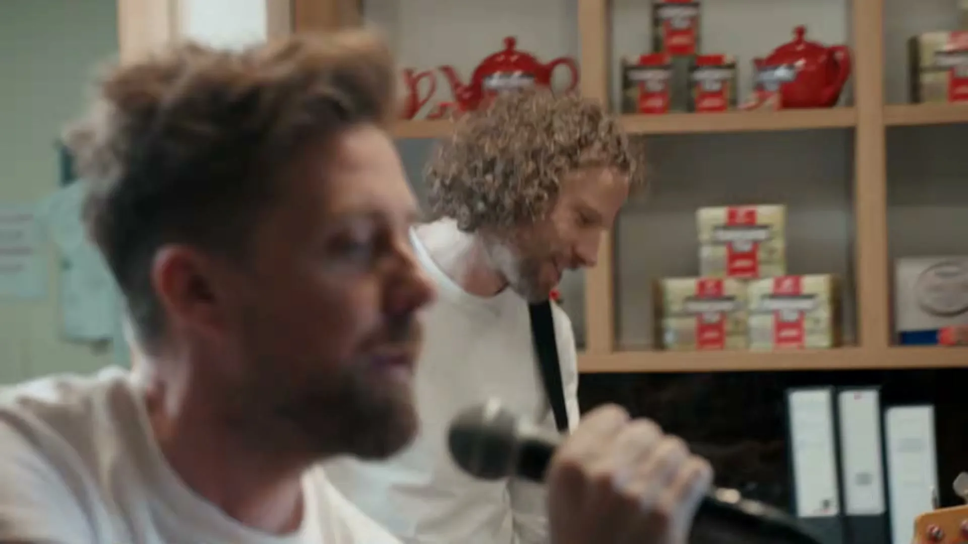 Two men, one with a microphone and the other with a guitar, perform in an office with shelves of tea products in the background.