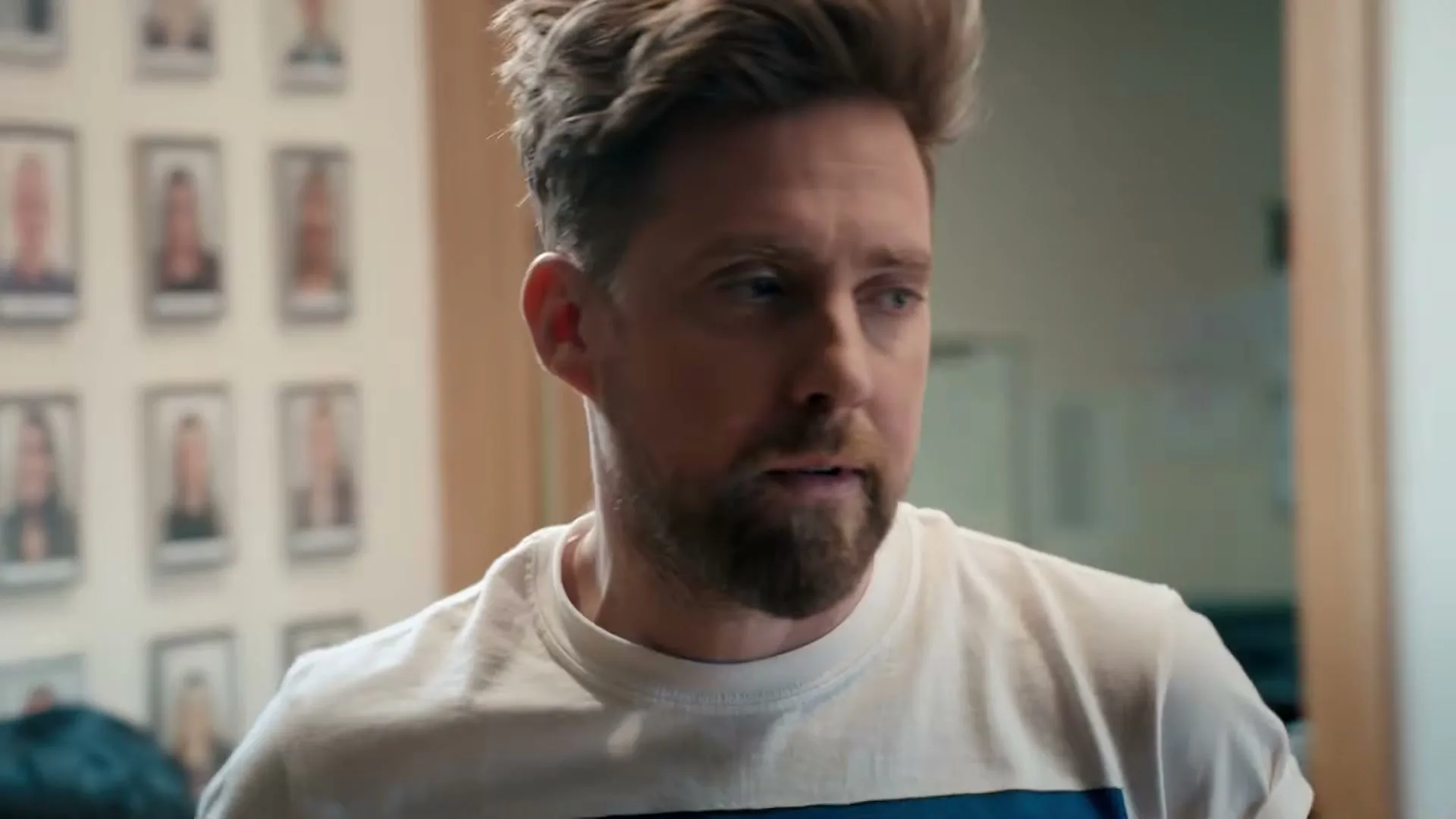 A man with a beard and a white t-shirt looks around a recording studio, with a microphone in the foreground.